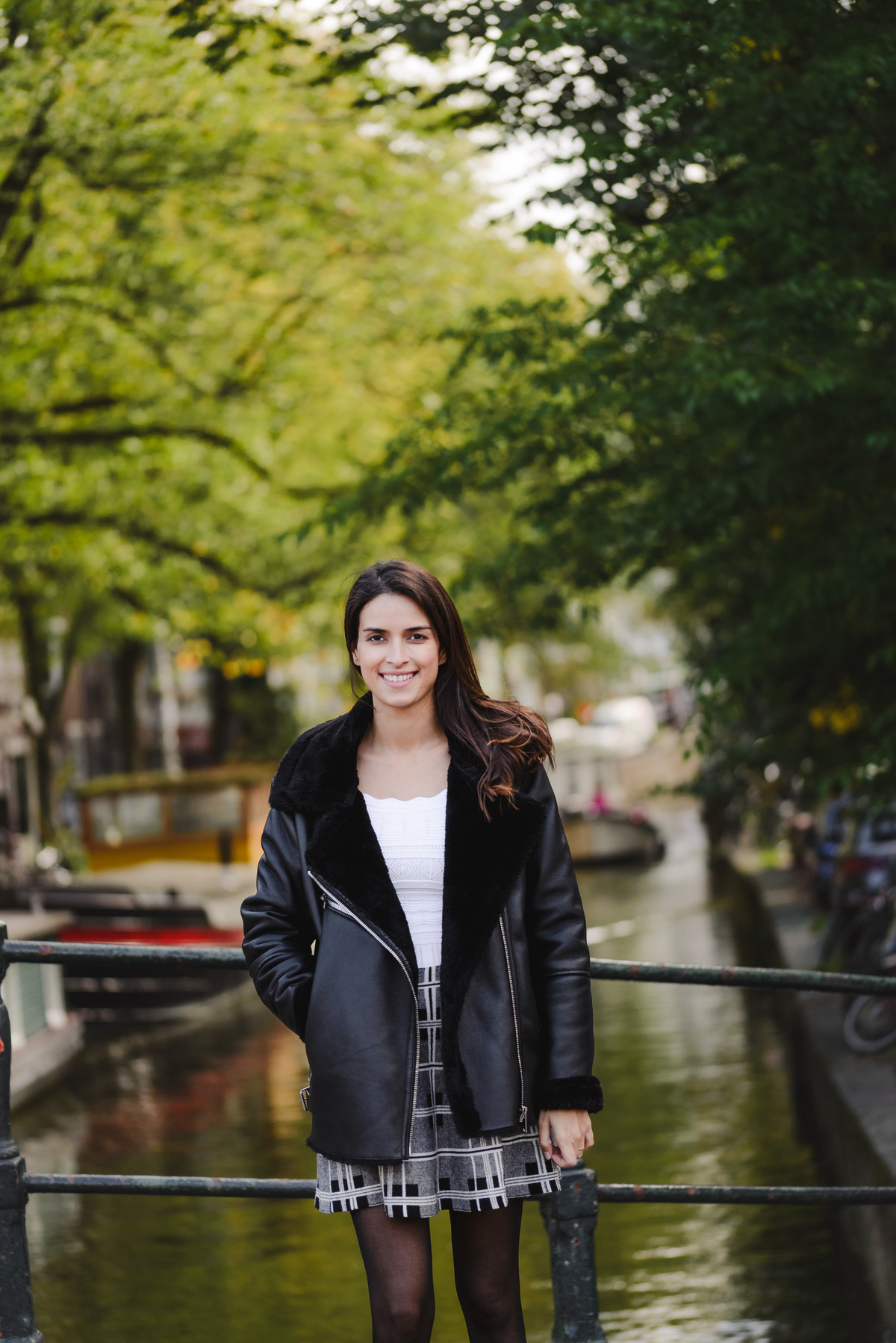 Woman standing on a bridge, smiling toward the camera, with houseboats and a green canal behind her.