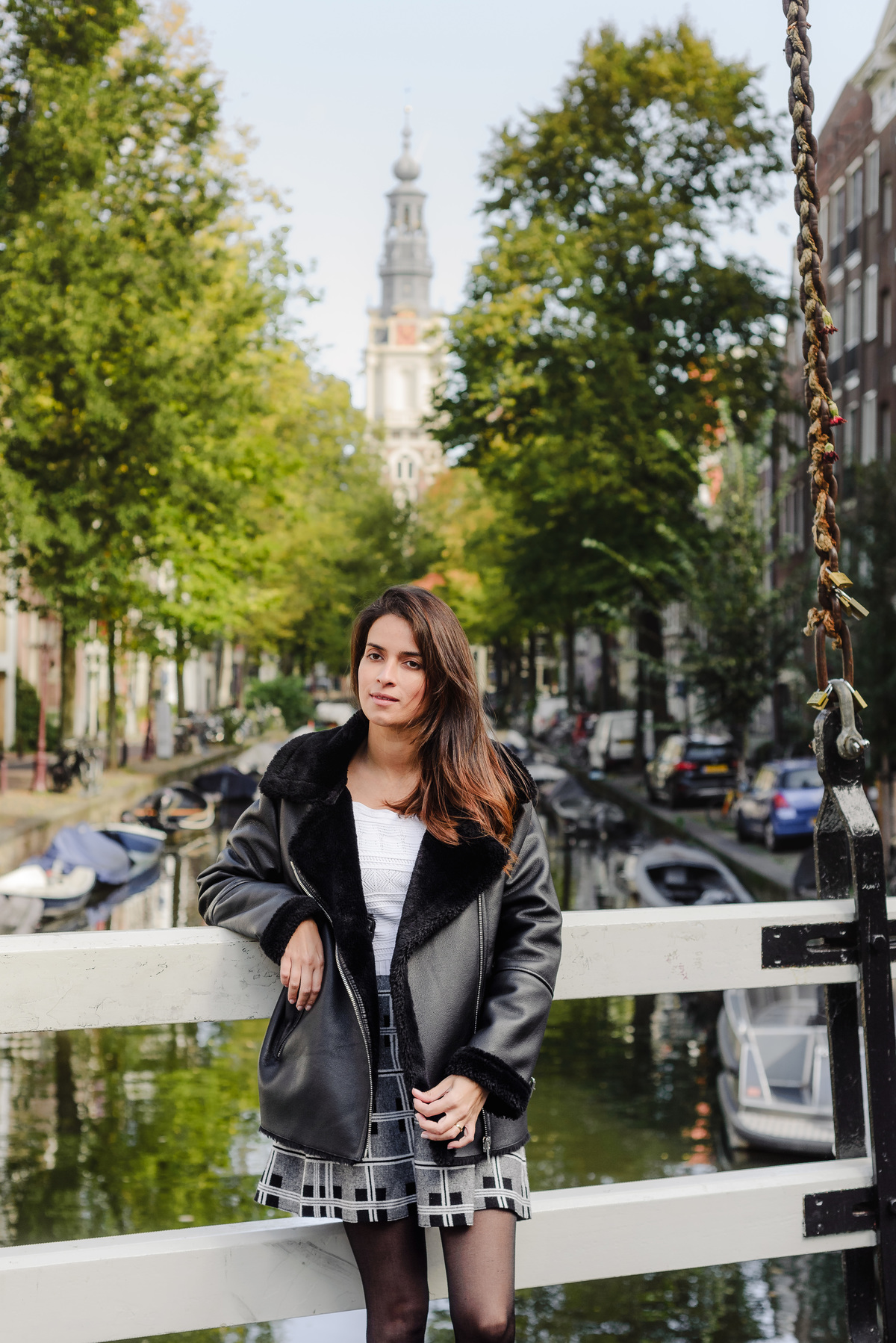 Woman standing on a canal bridge in Amsterdam, with green trees and a historic church tower in the background.