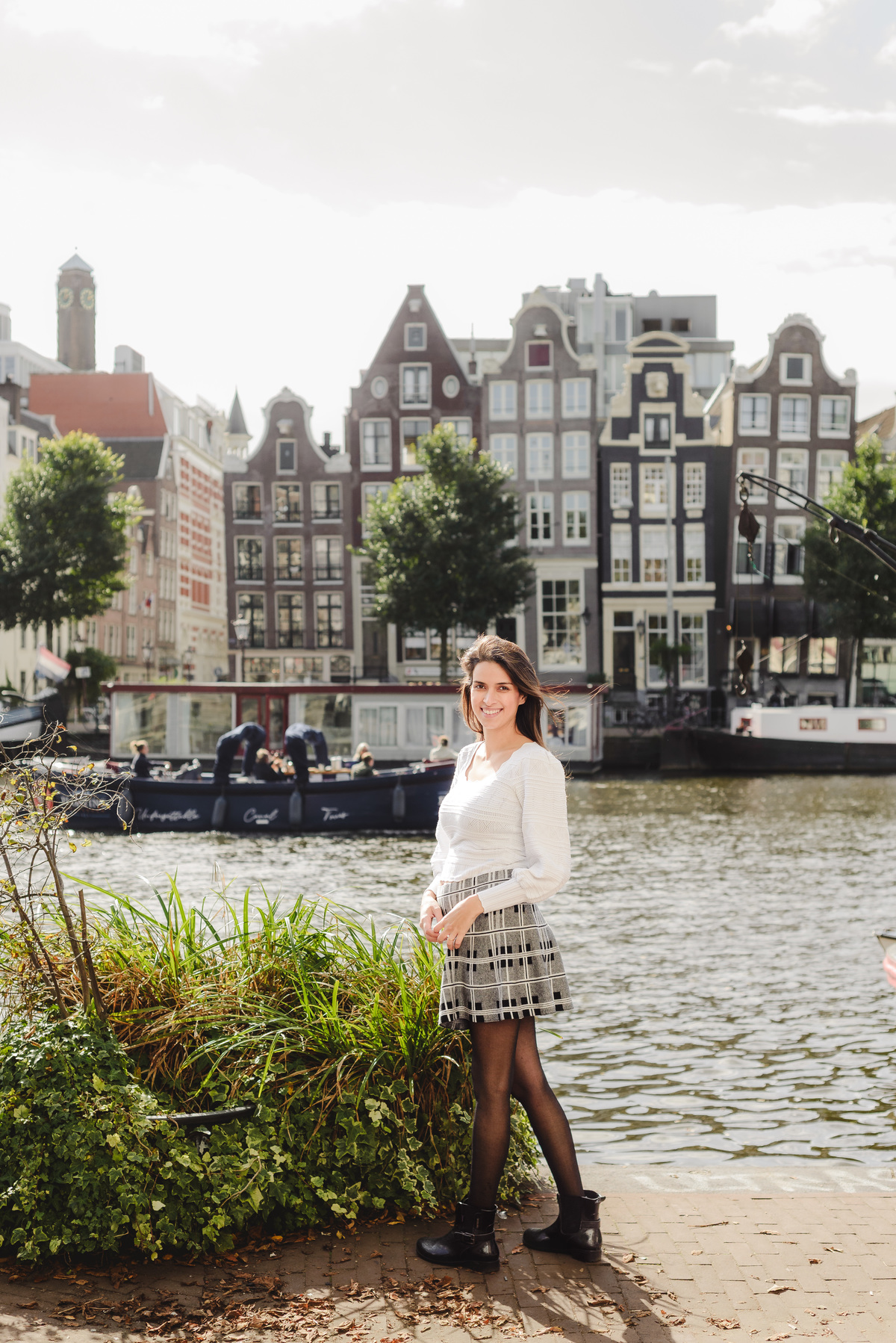 Woman posing by the water with Amsterdam’s canal houses behind her, hands resting on her belly.
