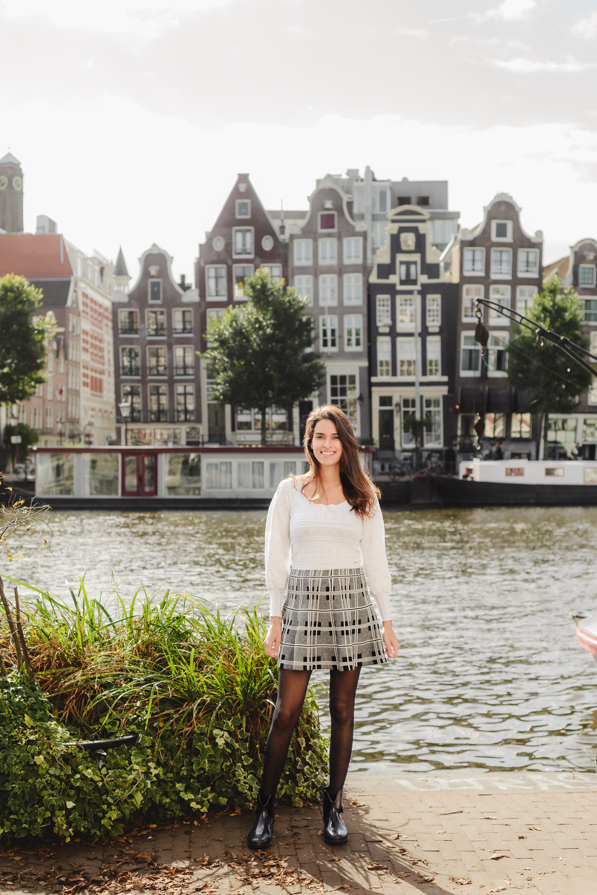 Woman standing by a canal with sunlight on her face, framed by historic Amsterdam houses.