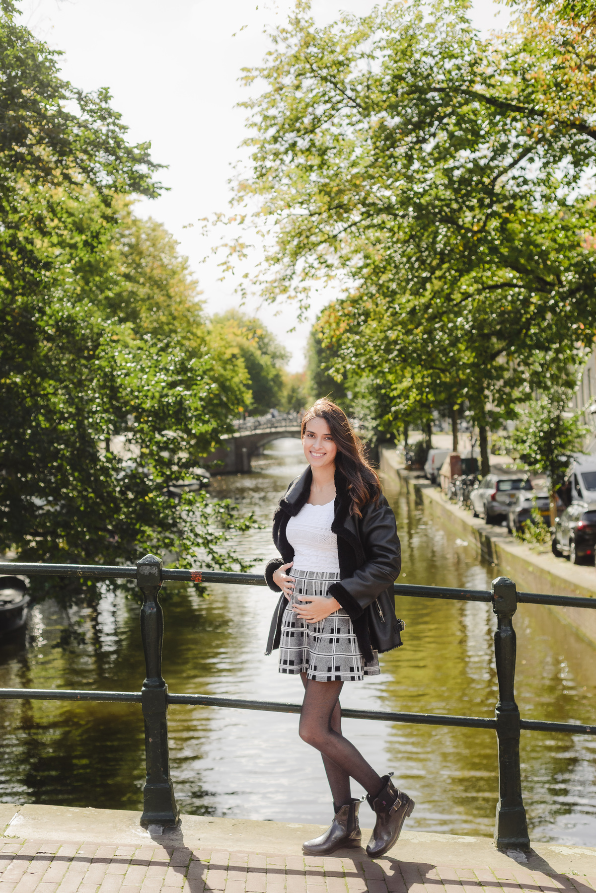 Woman on a bridge over a quiet Amsterdam canal, holding her belly and smiling.