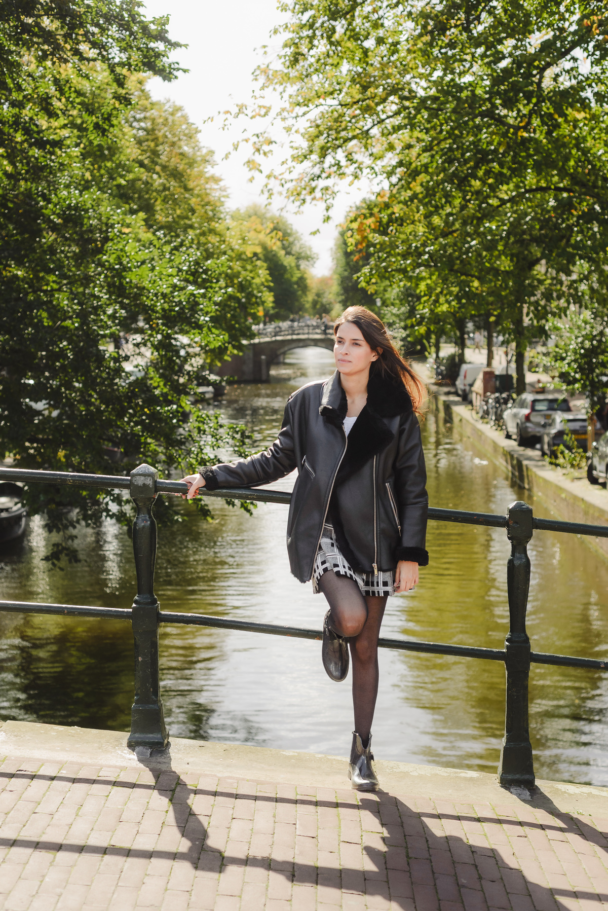 Woman stands on a canal bridge in Amsterdam, leaning on the railing with one leg raised, wearing a black jacket and plaid skirt, with trees and a distant bridge behind her.