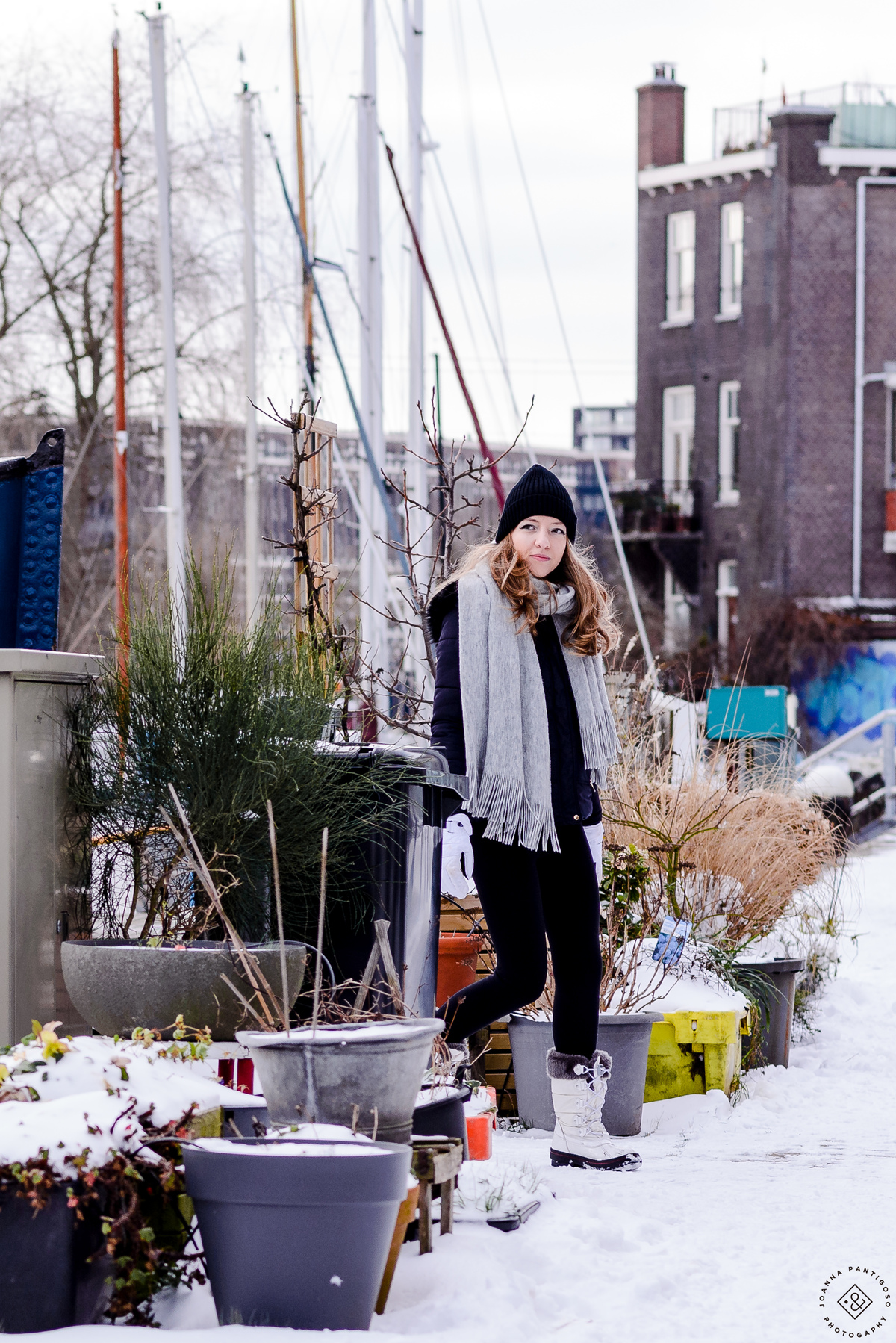 Woman standing near houseboats in Amsterdam during snowfall, dressed in winter boots, scarf, and hat, surrounded by snow-covered plants and pavement.