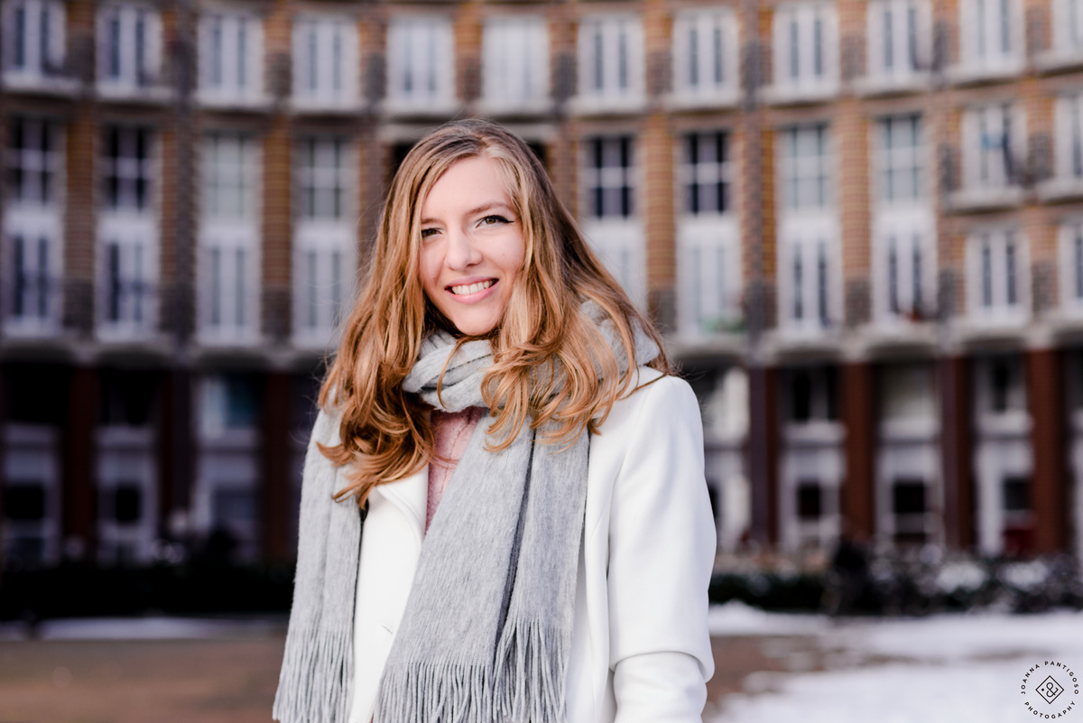Portrait of a woman smiling in winter in Amsterdam, wearing a light coat and scarf, with a brick residential building softly blurred in the background.