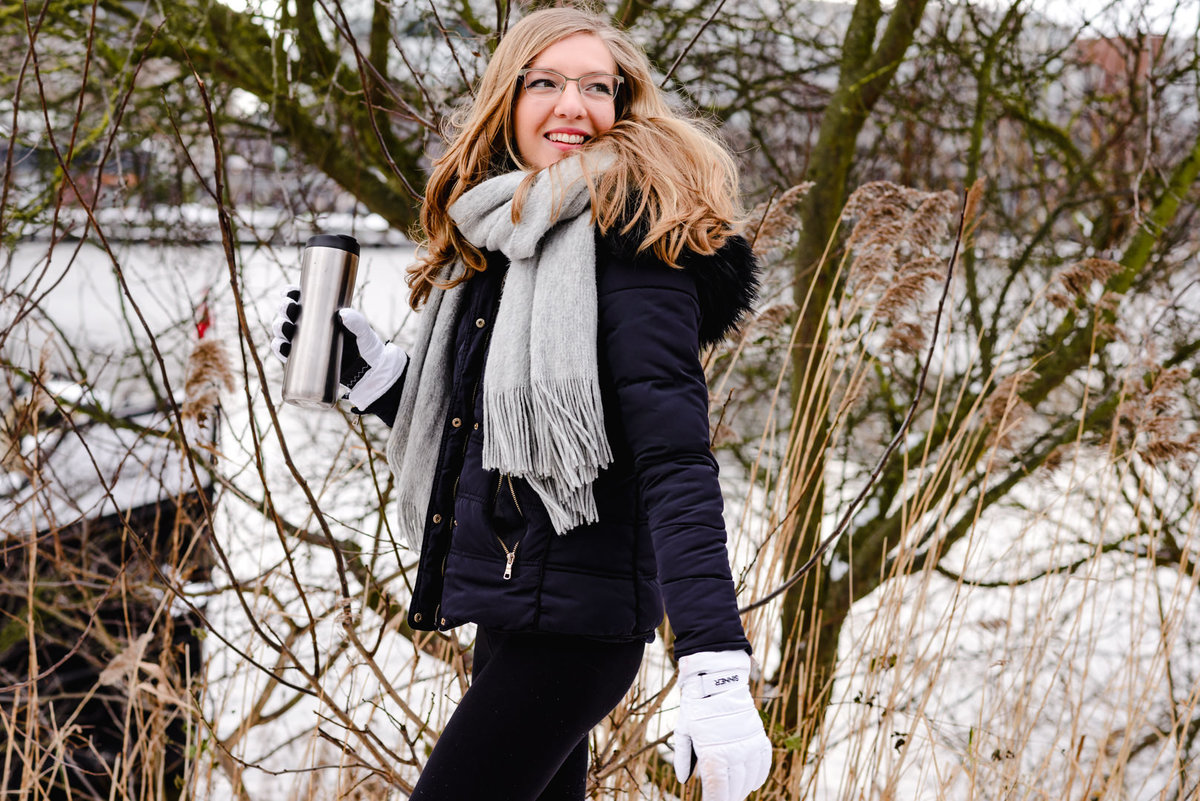 Woman walking outdoors in Amsterdam during winter, wearing a dark coat, scarf, and gloves, holding a thermos with snow-covered trees in the background.