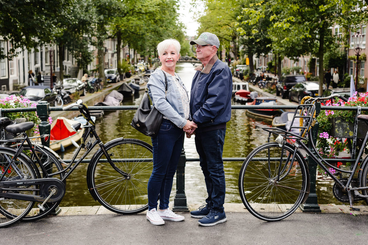 Couple holding hands on a bridge over a quiet Amsterdam canal, bicycles framing the scene and trees lining the water.