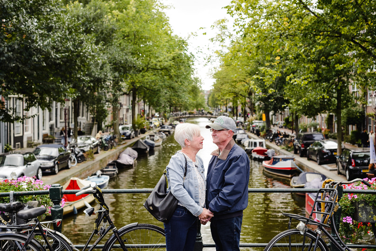 Couple standing close on a canal bridge, facing each other with boats and historic houses in the background.