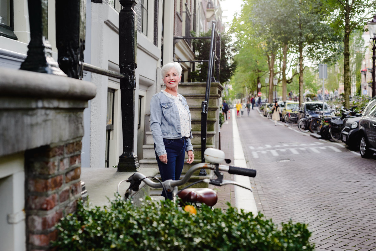 Woman standing alone on a residential Amsterdam street near canal houses, bicycles and pedestrians in the background.