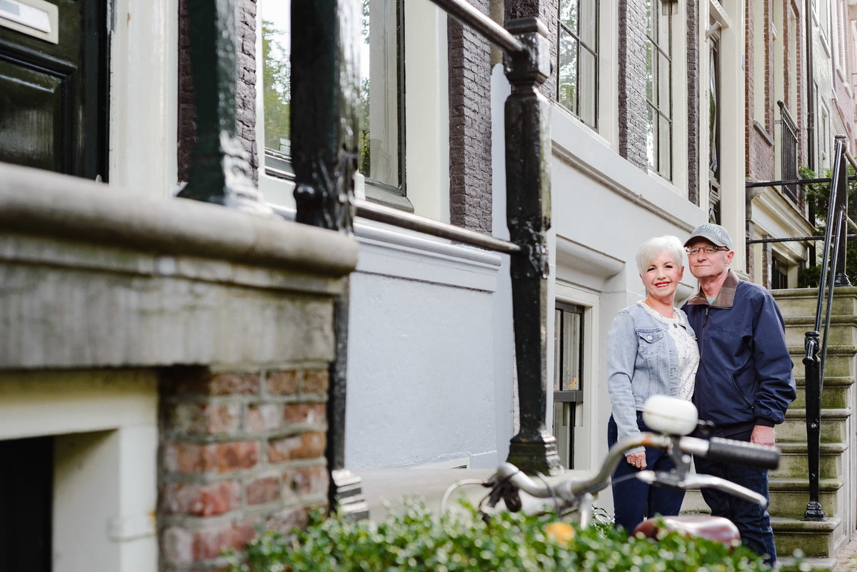 Couple standing together near the steps of a canal house, framed by railings and greenery.