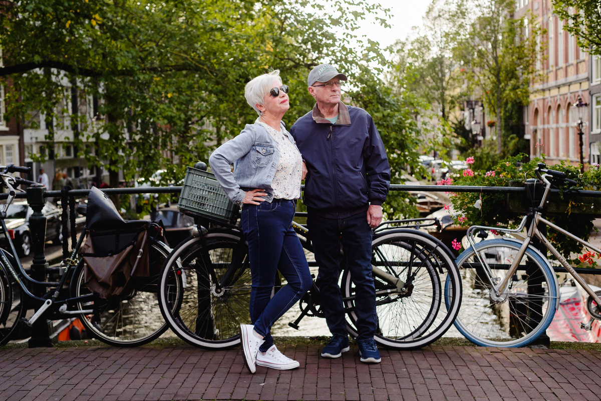 Mature couple standing on a canal bridge in Amsterdam, bicycles beside them, looking off into the distance among green trees.