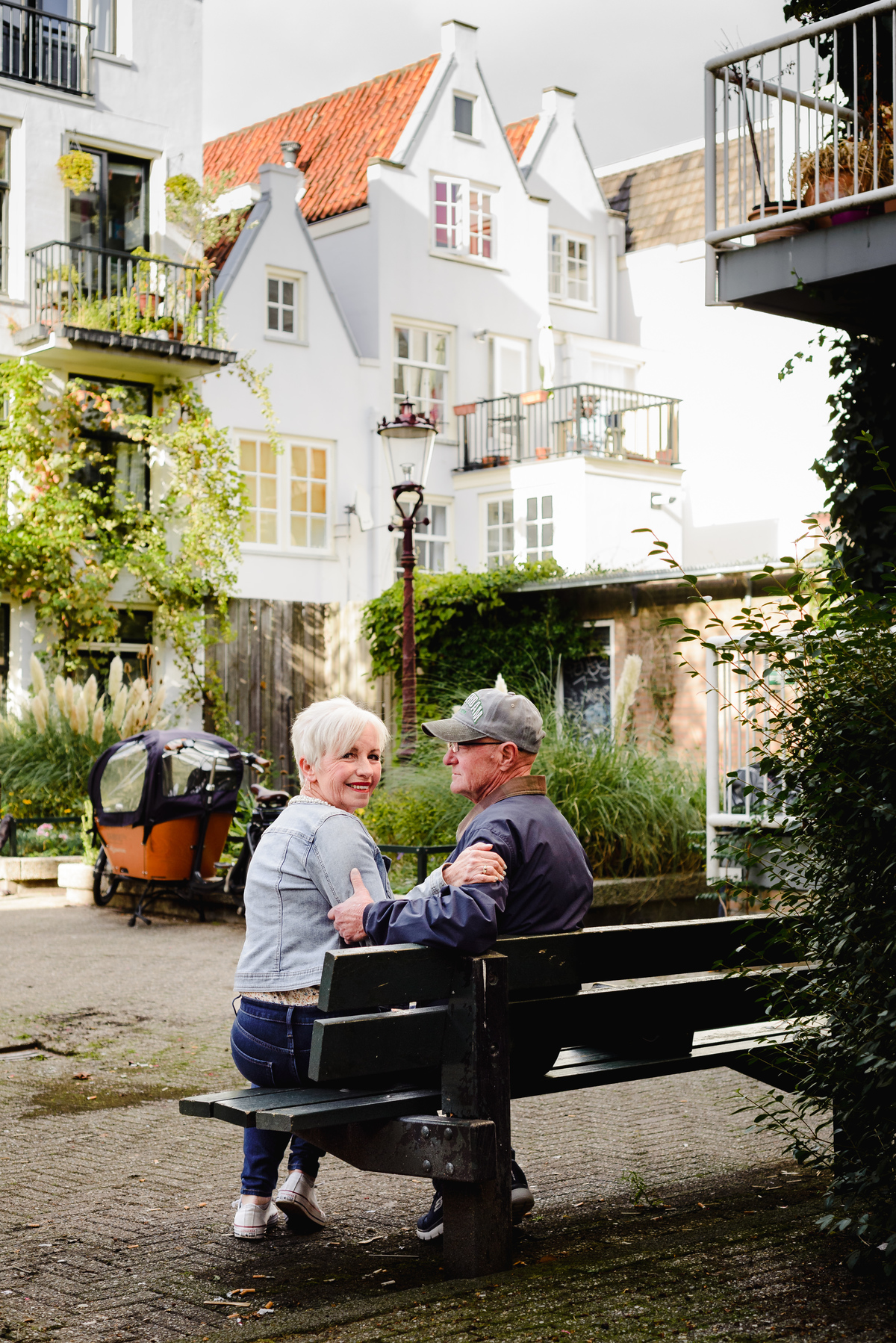 Couple sitting close together on a bench in a quiet Amsterdam courtyard with white houses and greenery around them.
