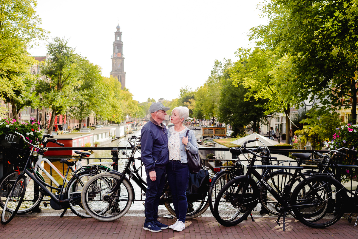 Couple standing together on an Amsterdam canal bridge with the Westertoren visible in the background and boats on the water.