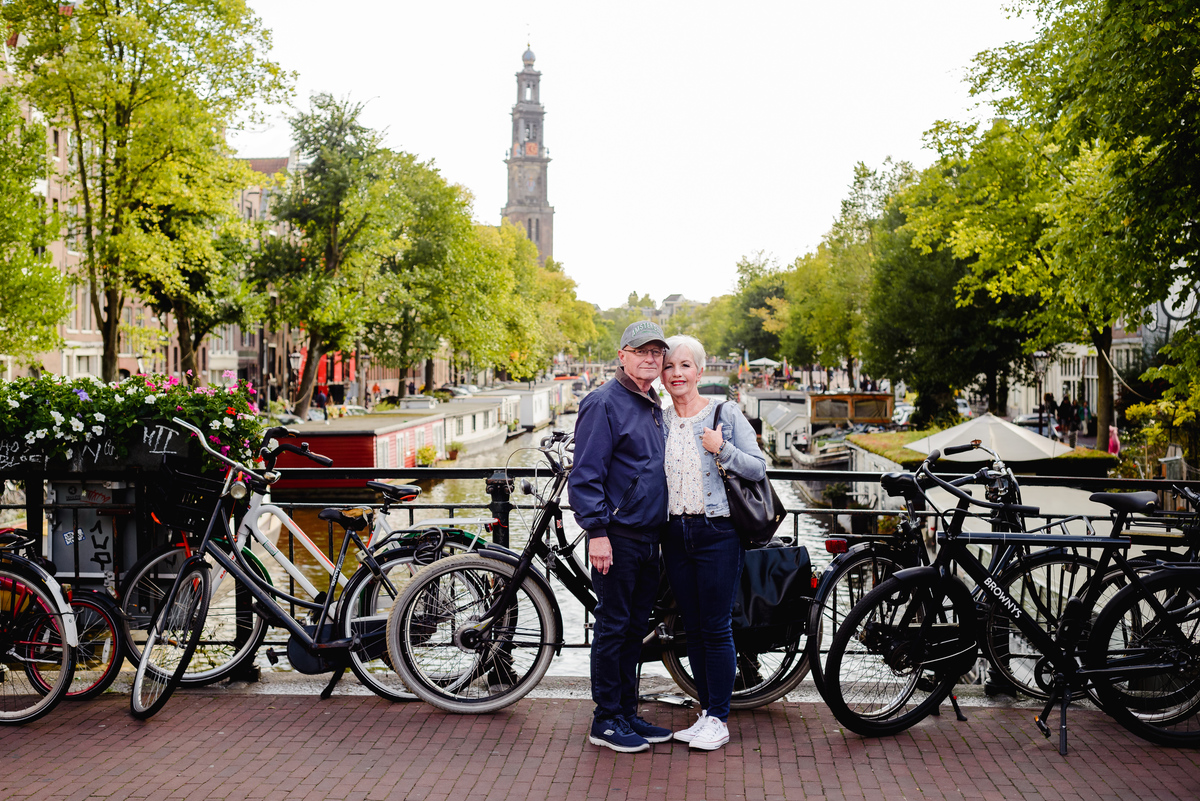 Anniversary couple posing on a canal bridge in Amsterdam, framed by bicycles, houseboats, and tree-lined canals.