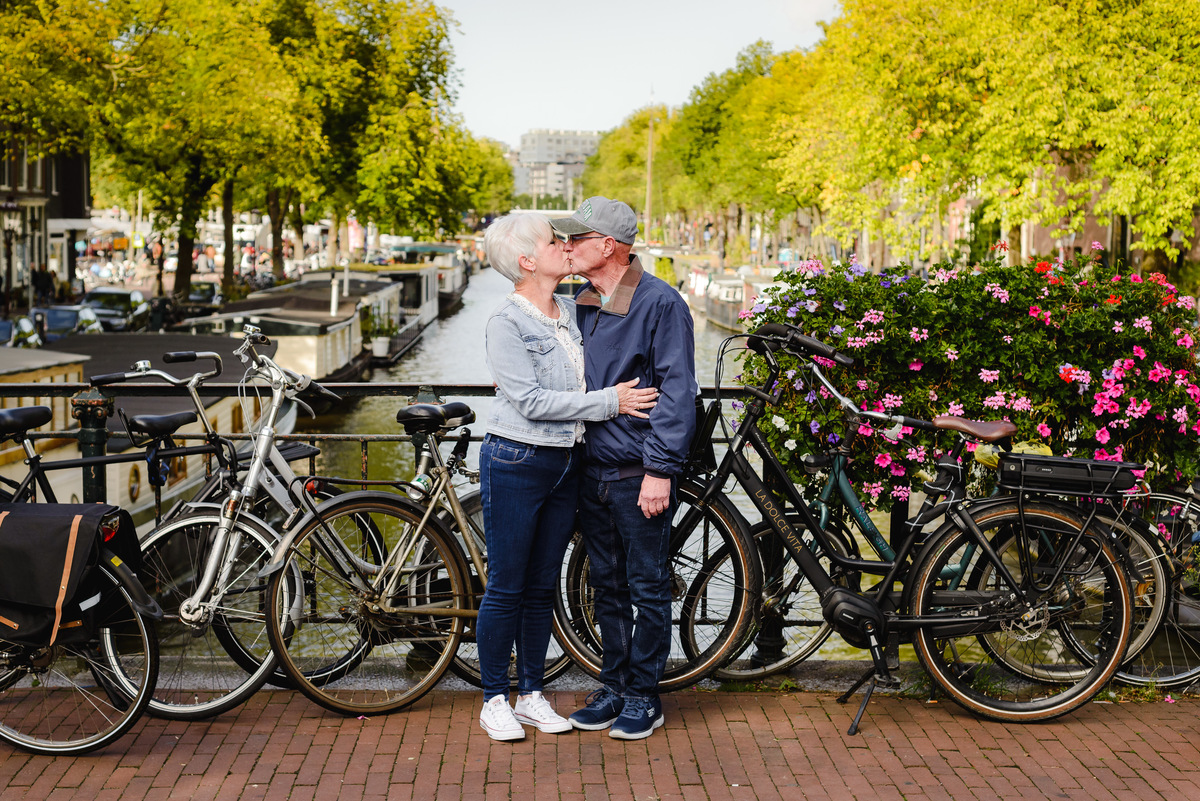 Romantic kiss between a couple on an Amsterdam canal bridge, bicycles and colorful flowers lining the railing.