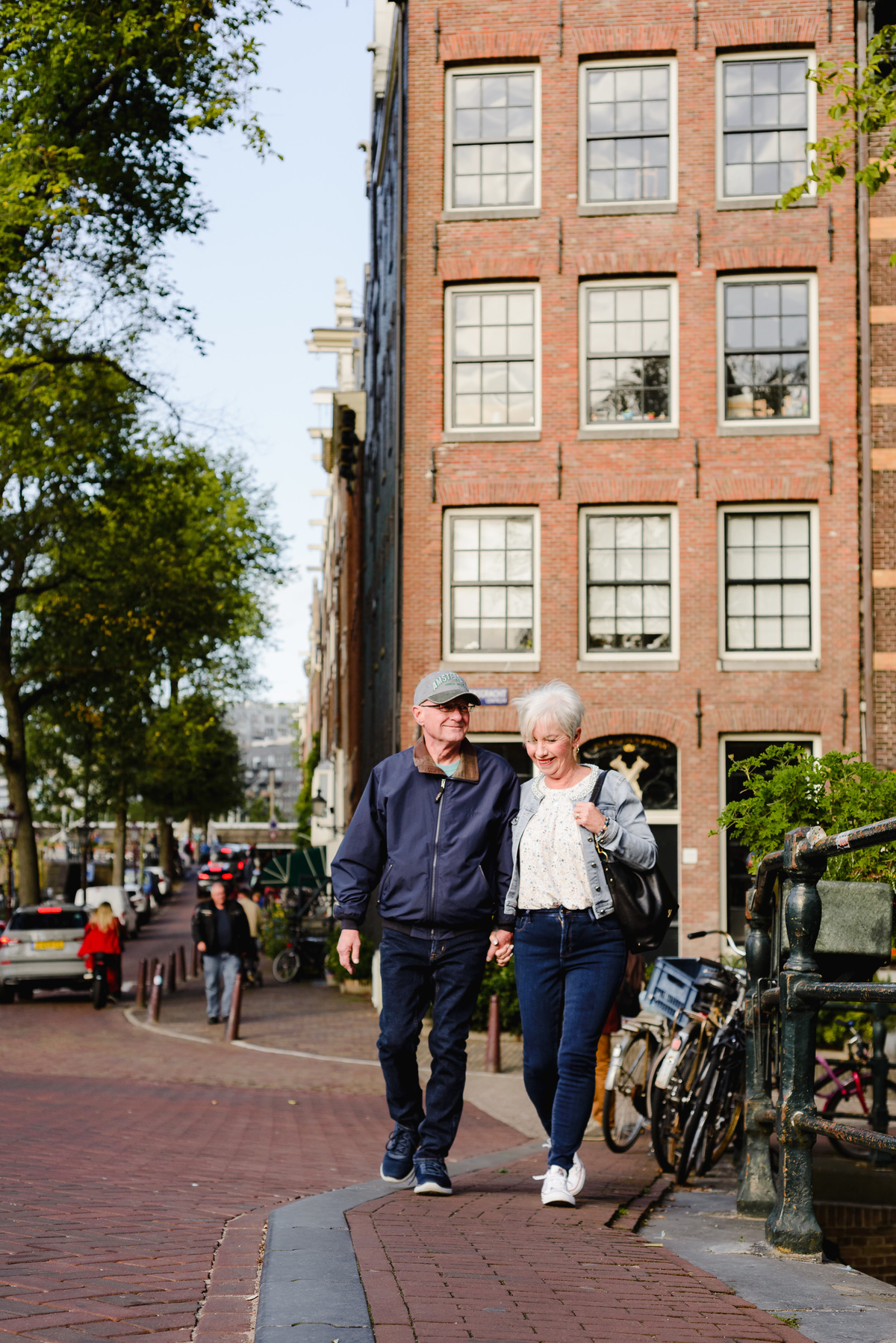 Couple walking hand in hand along a quiet Amsterdam street during an anniversary photo session, brick buildings and trees in the background.