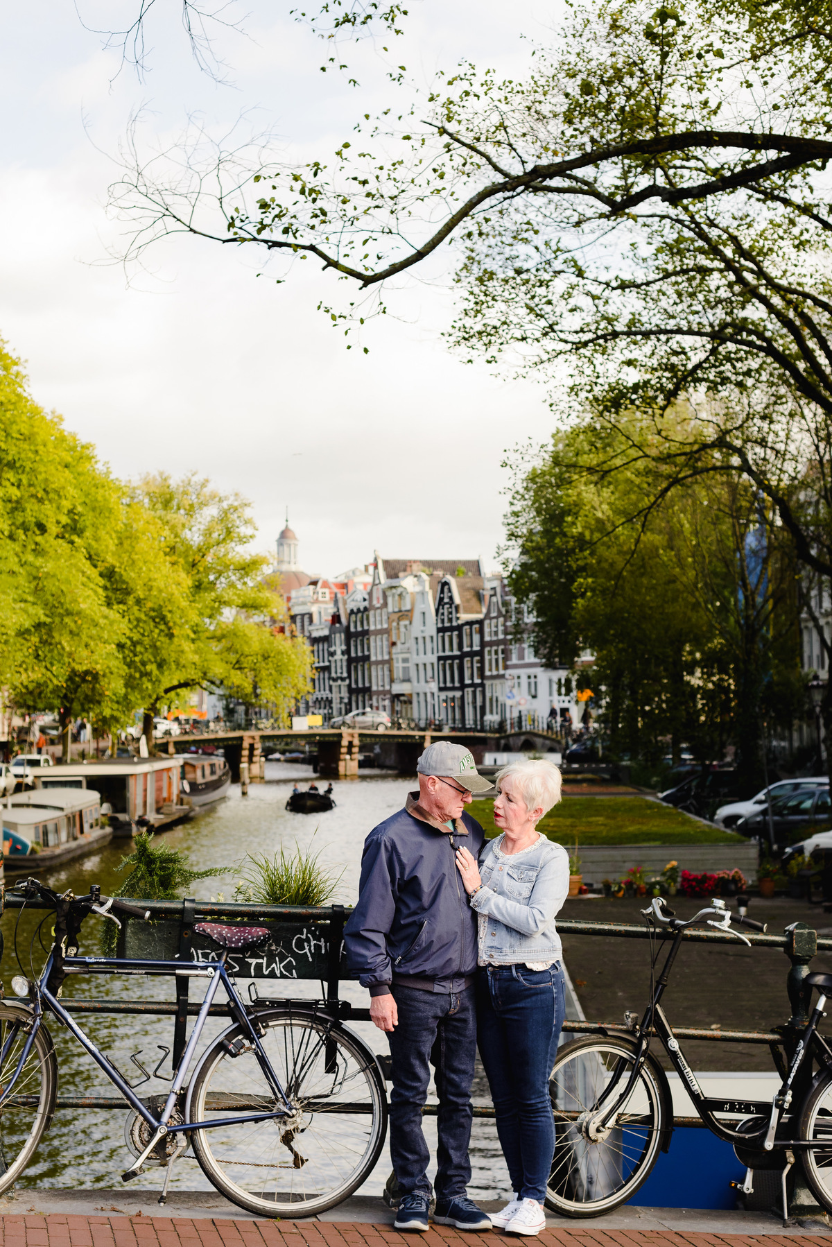 Anniversary couple standing on a small Amsterdam bridge, looking at each other with canals and historic houses behind them.