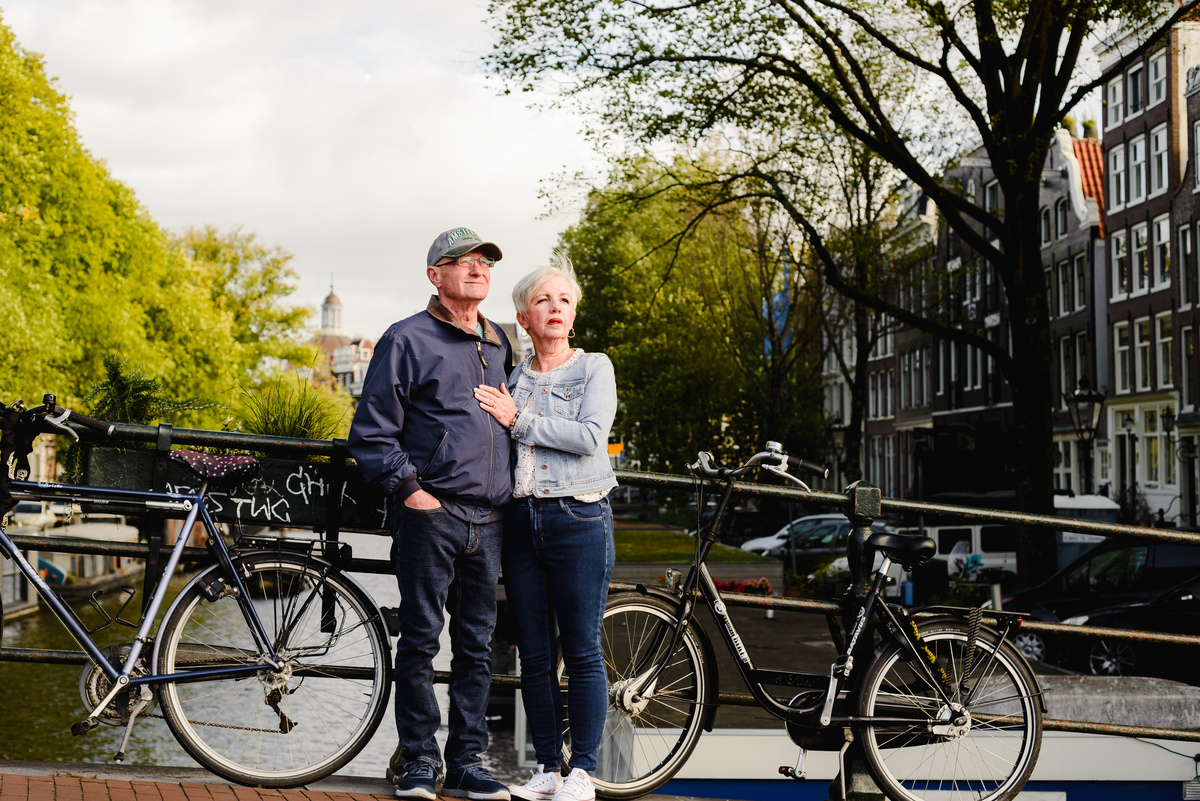 Older couple posing together on a canal bridge in Amsterdam, framed by trees, bicycles, and classic canal houses.