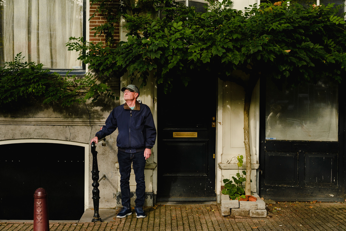 Portrait of a man standing under leafy shadows by a traditional Amsterdam canal house facade.
