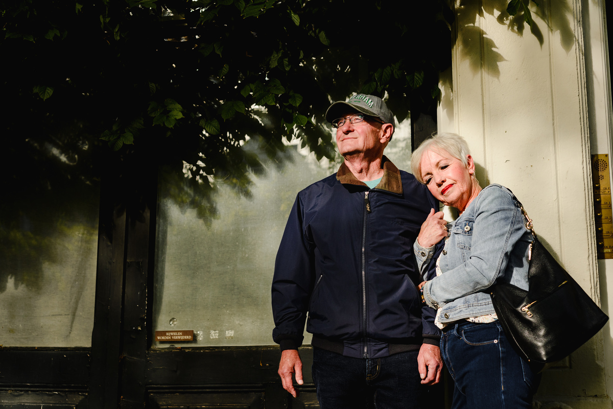 Couple leaning together under tree shadows beside a canal house in Amsterdam, relaxed and intimate moment.