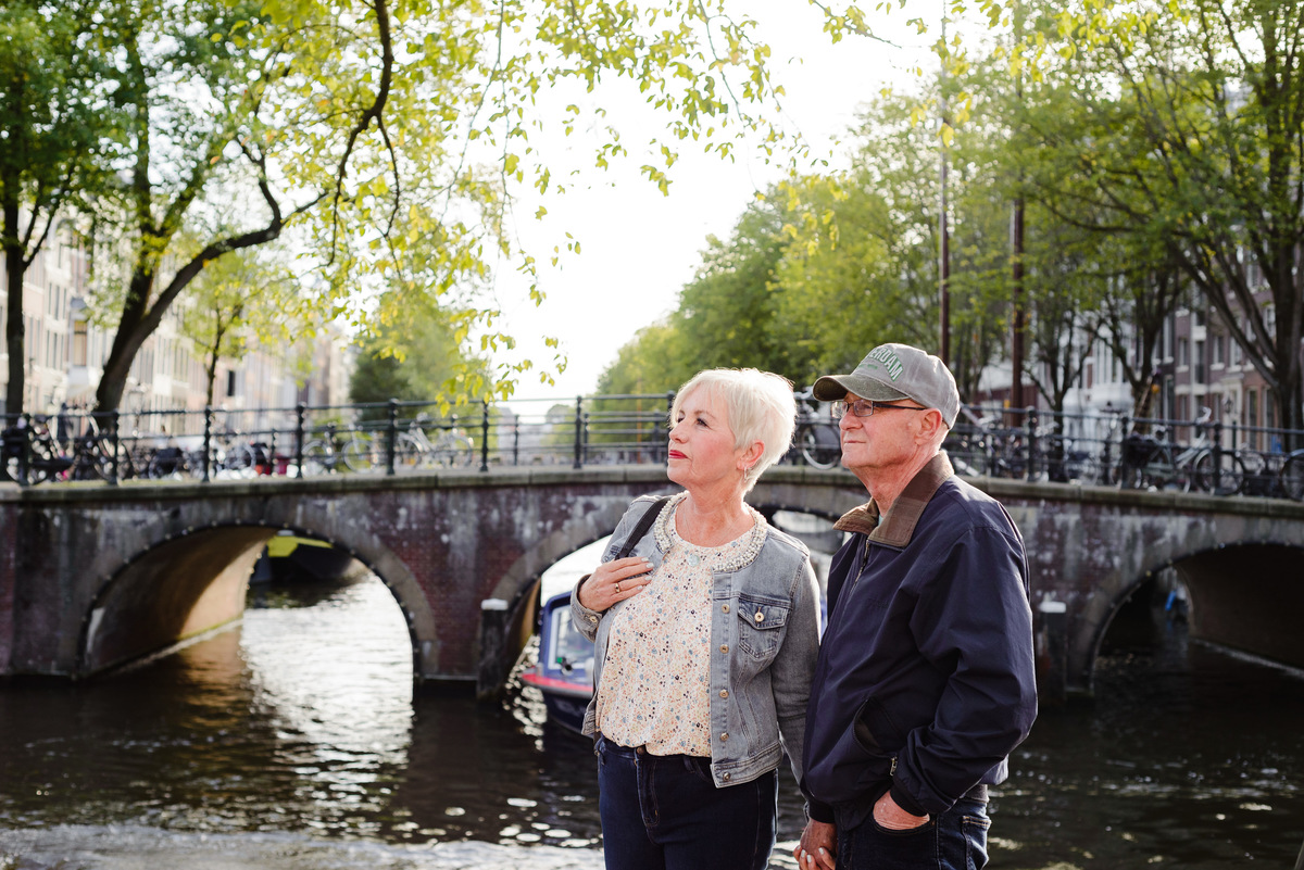 Anniversary couple standing by an Amsterdam canal with an arched bridge in the background, looking into the distance.