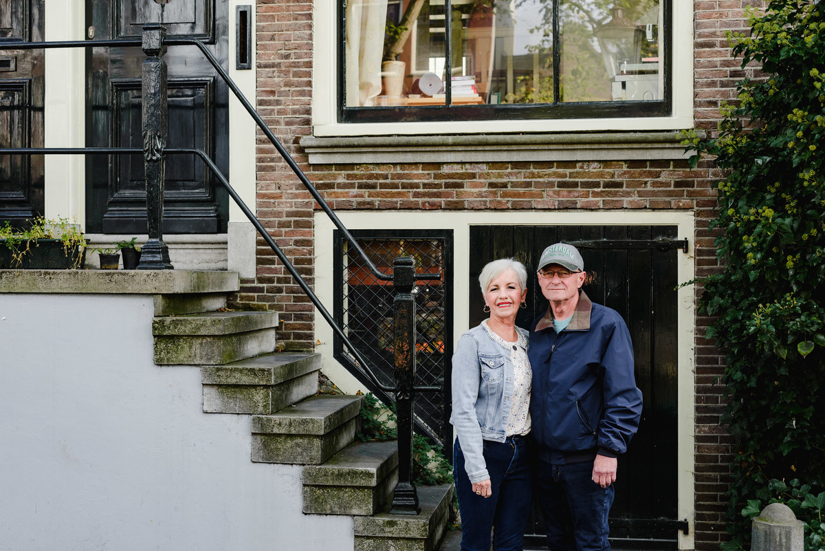 Anniversary couple posing in front of a traditional Amsterdam canal house with brick façade and large window.