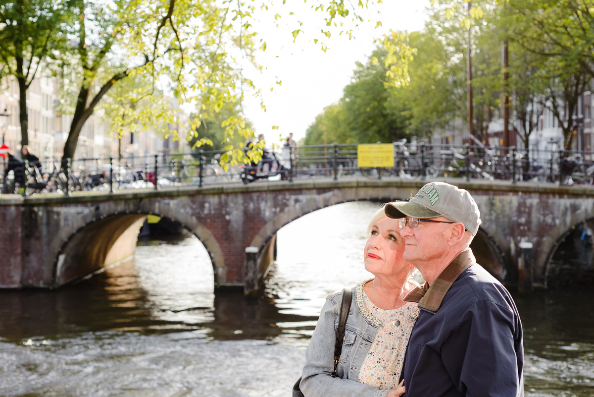 Close portrait of a couple by an Amsterdam canal, sunlight reflecting on the water and brick bridge behind them.