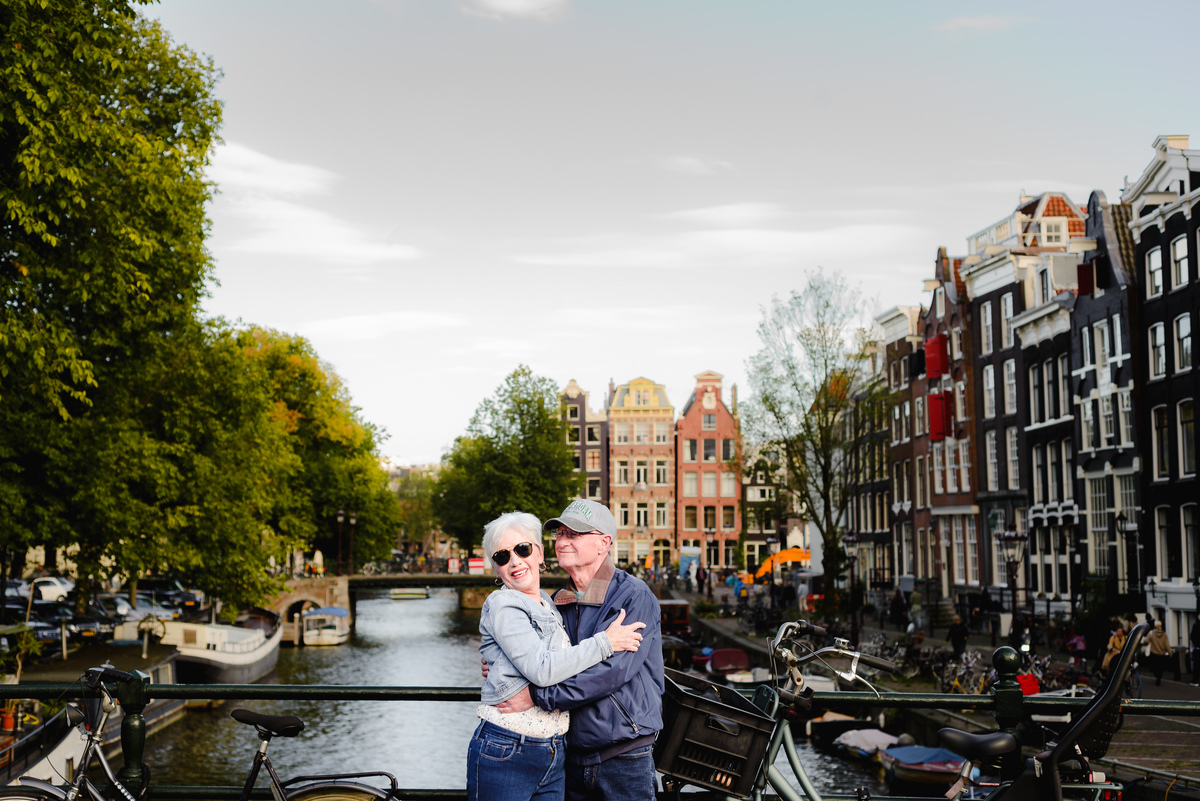 Older couple embracing on a canal bridge in Amsterdam, smiling and holding each other with the city and water behind them.