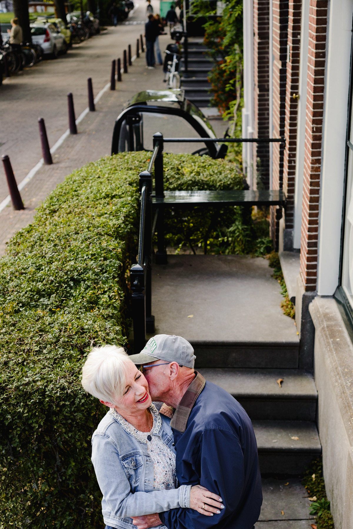 Close, affectionate portrait of an older couple hugging and smiling in a residential Amsterdam street, surrounded by greenery and brick houses.