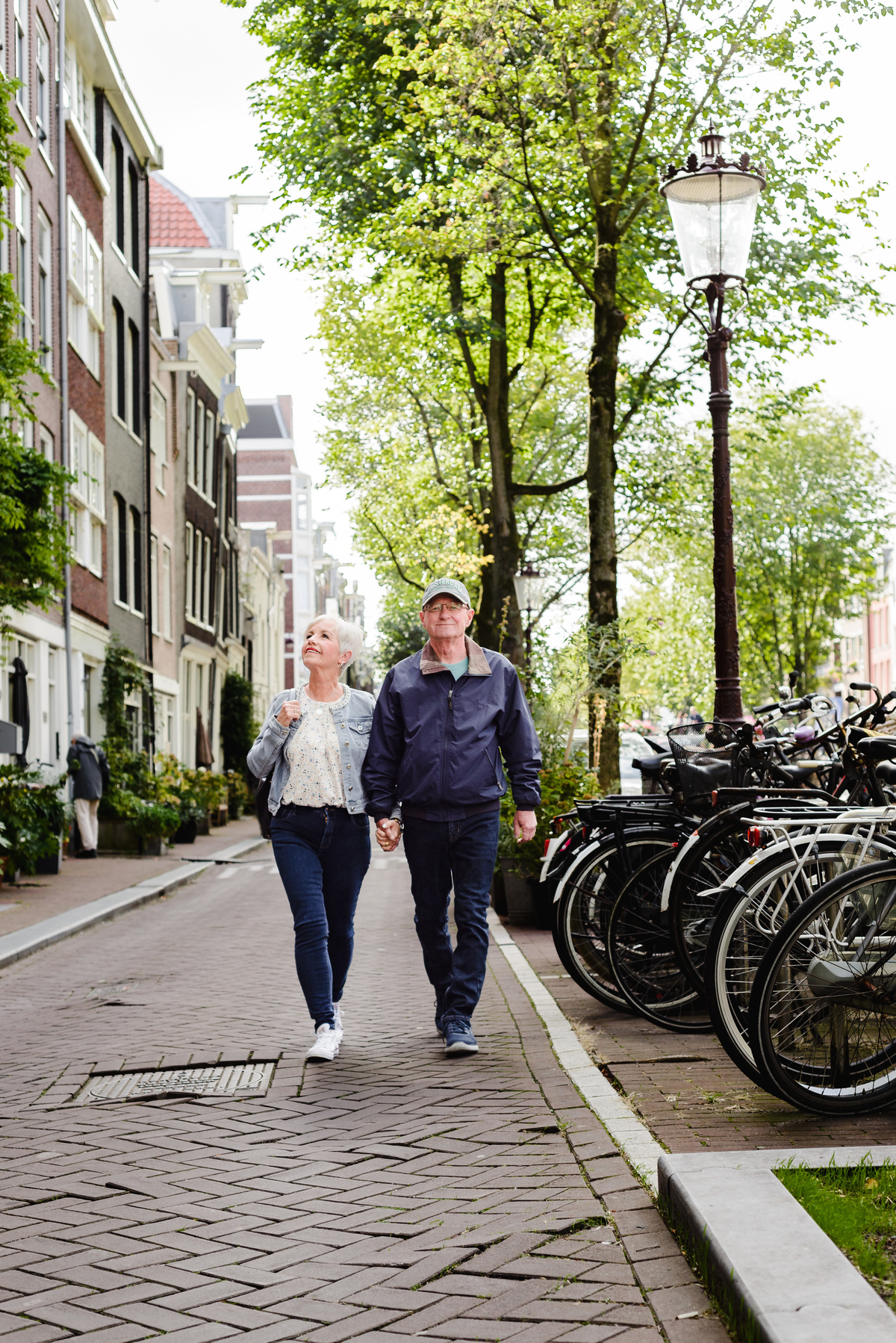 Anniversary couple photographed from a distance with soft foreground greenery, standing by a classic Amsterdam canal house entrance.
