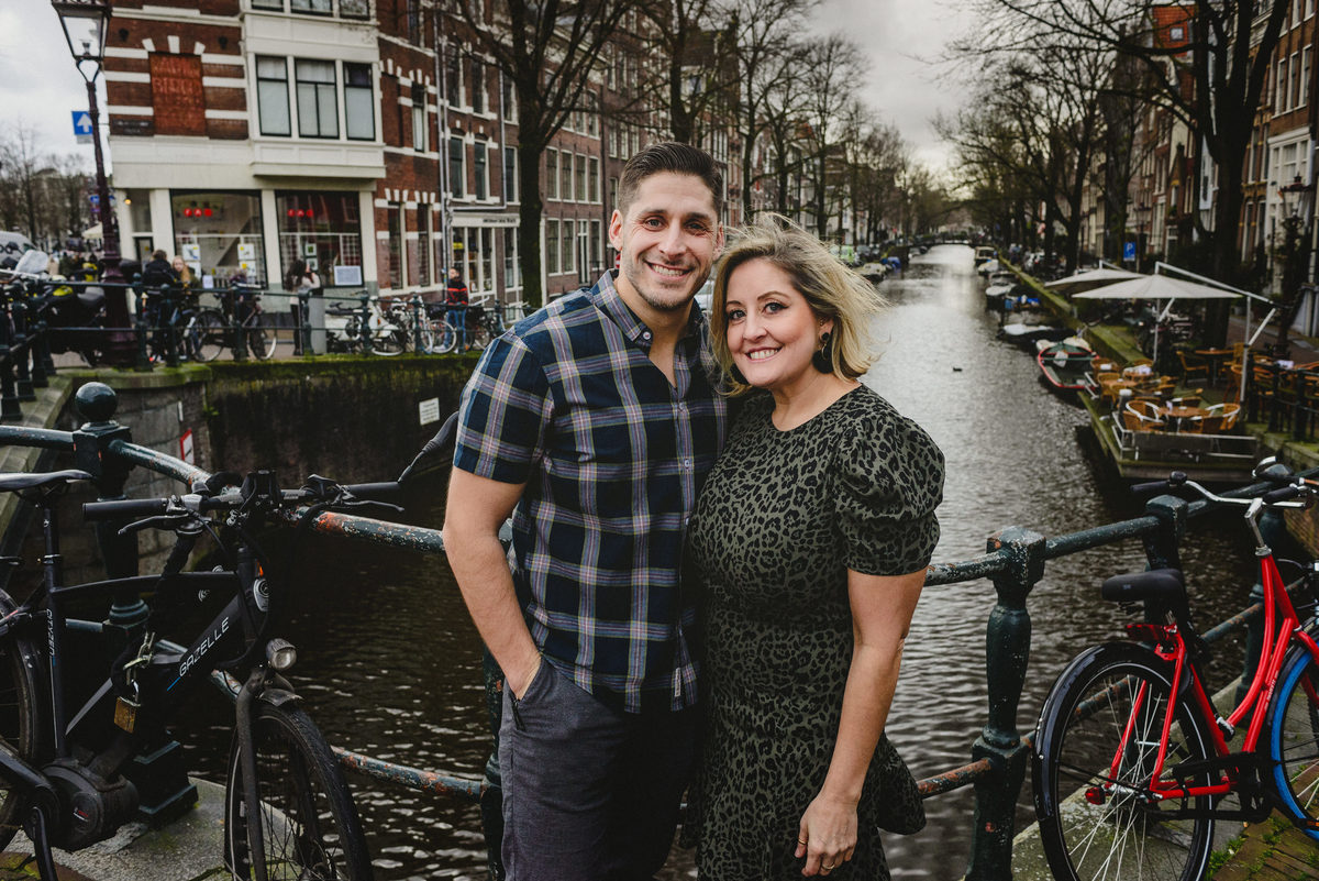 Adult siblings standing together on an Amsterdam canal bridge, smiling at the camera during a family photo session.