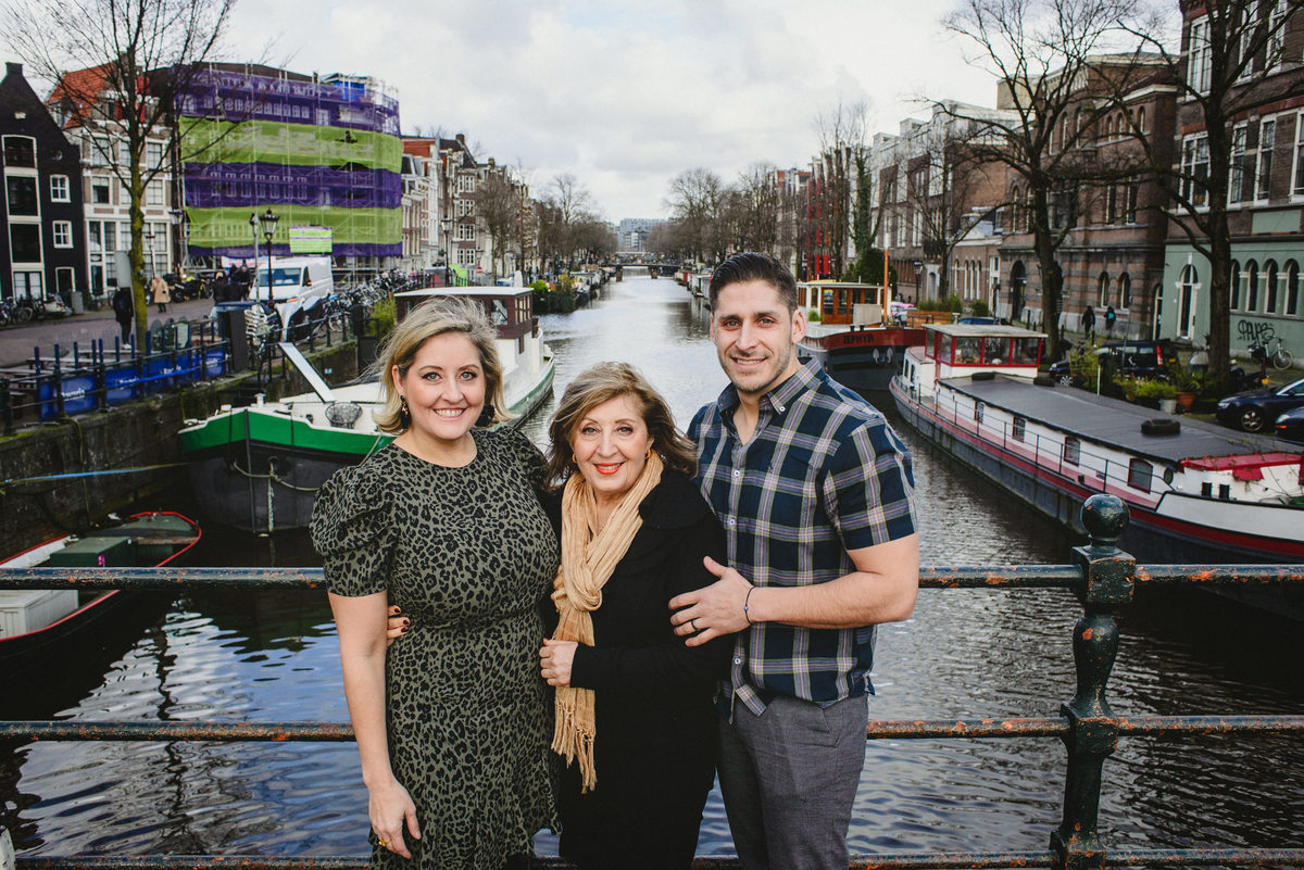Three adult family members standing close together on a canal bridge in Amsterdam, smiling at the camera, with houseboats and canal behind them.