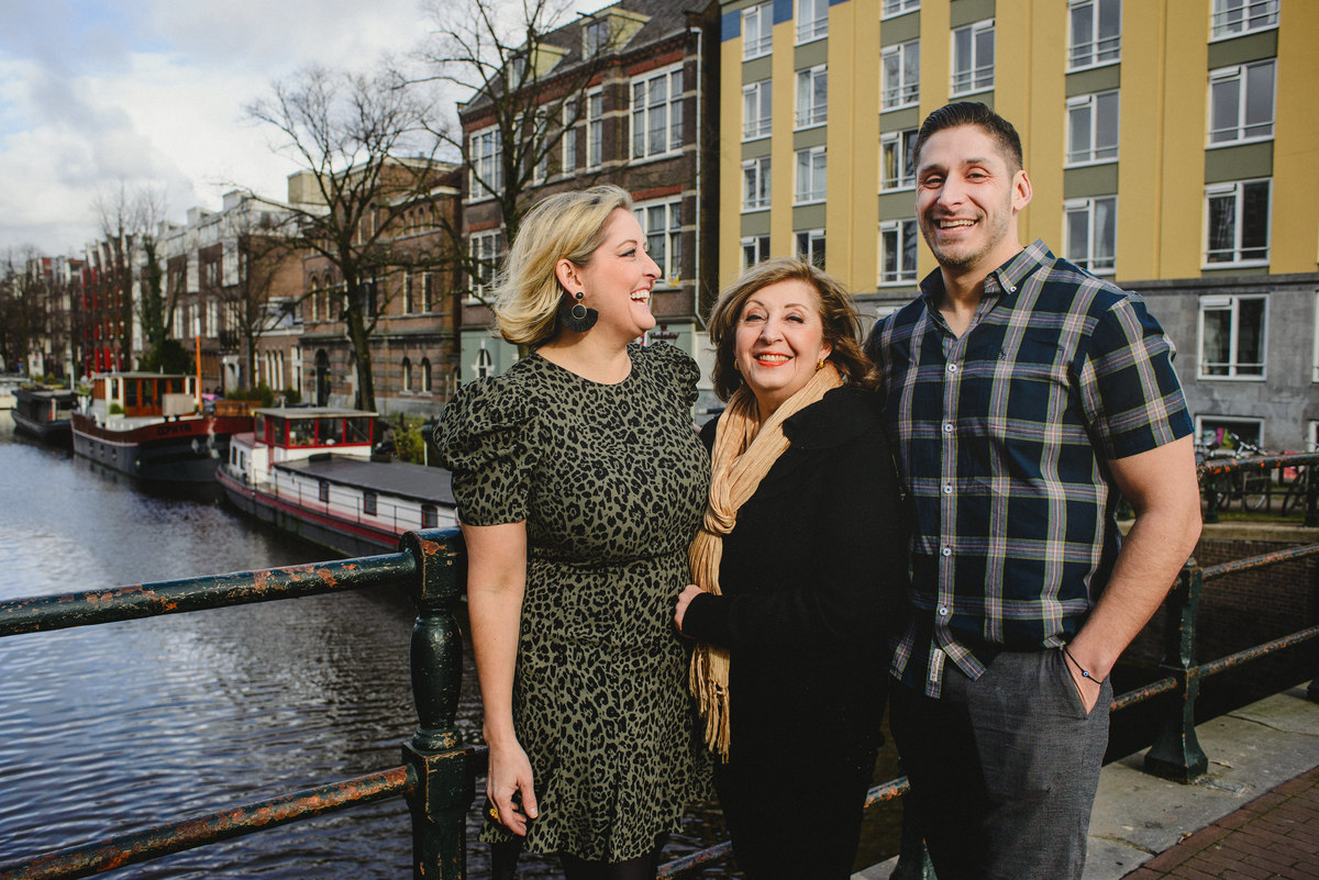 Family portrait of two adult children and their mother on an Amsterdam canal bridge, winter light and boats visible behind them.
