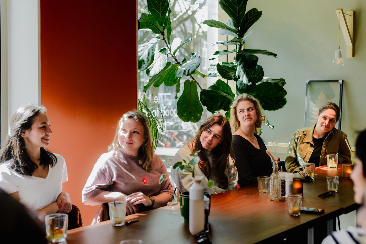 Women seated at a long table during an Amsterdam International Women welcome brunch, listening and smiling in a sunlit café.