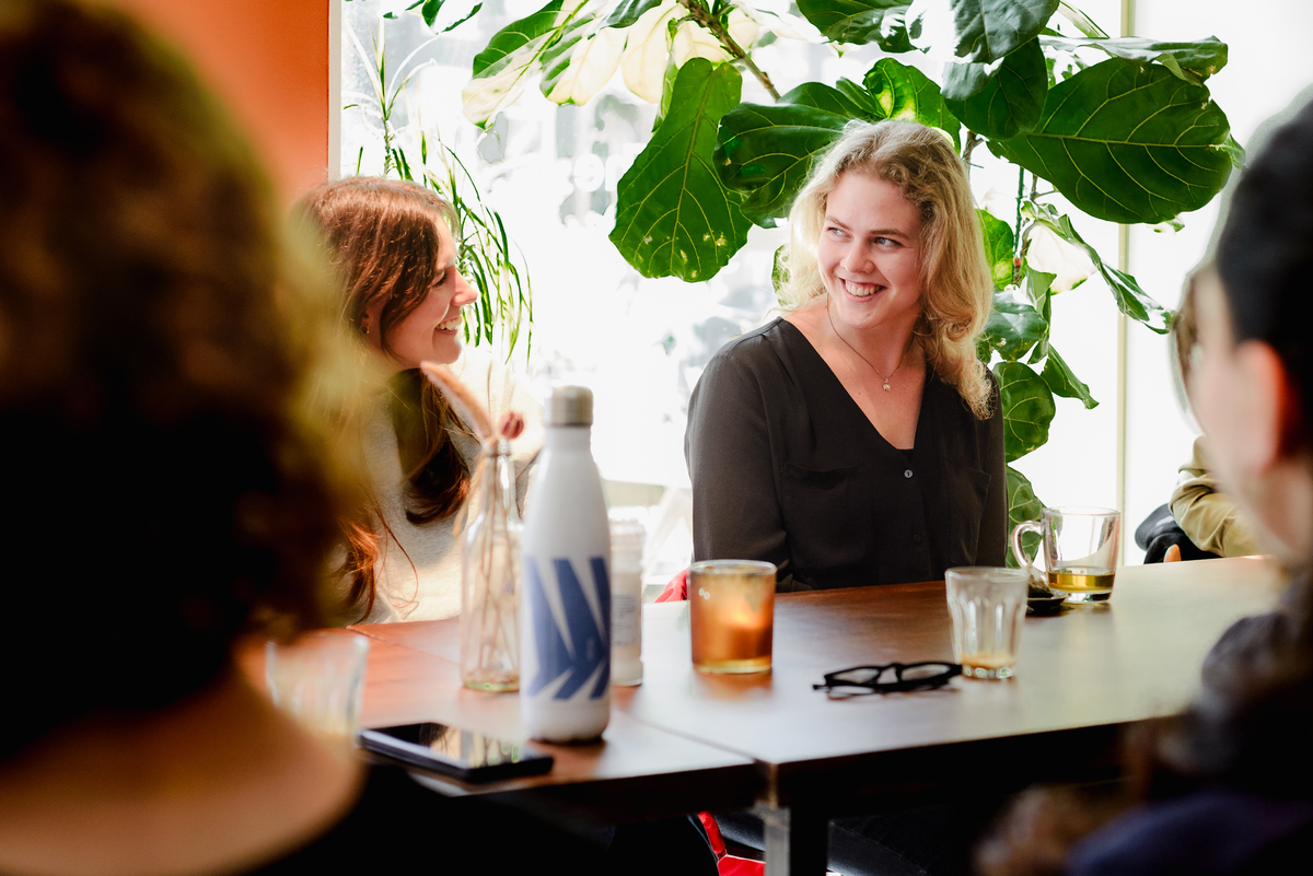 Two women smiling and talking at a community brunch event in Amsterdam, natural light and indoor plants in the background.