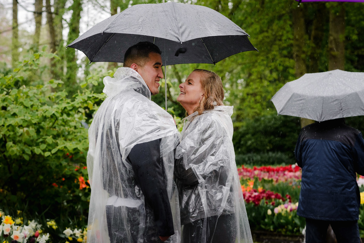 Engaged couple smiling at each other under an umbrella in Keukenhof gardens during light rain, spring flowers and trees in the background.