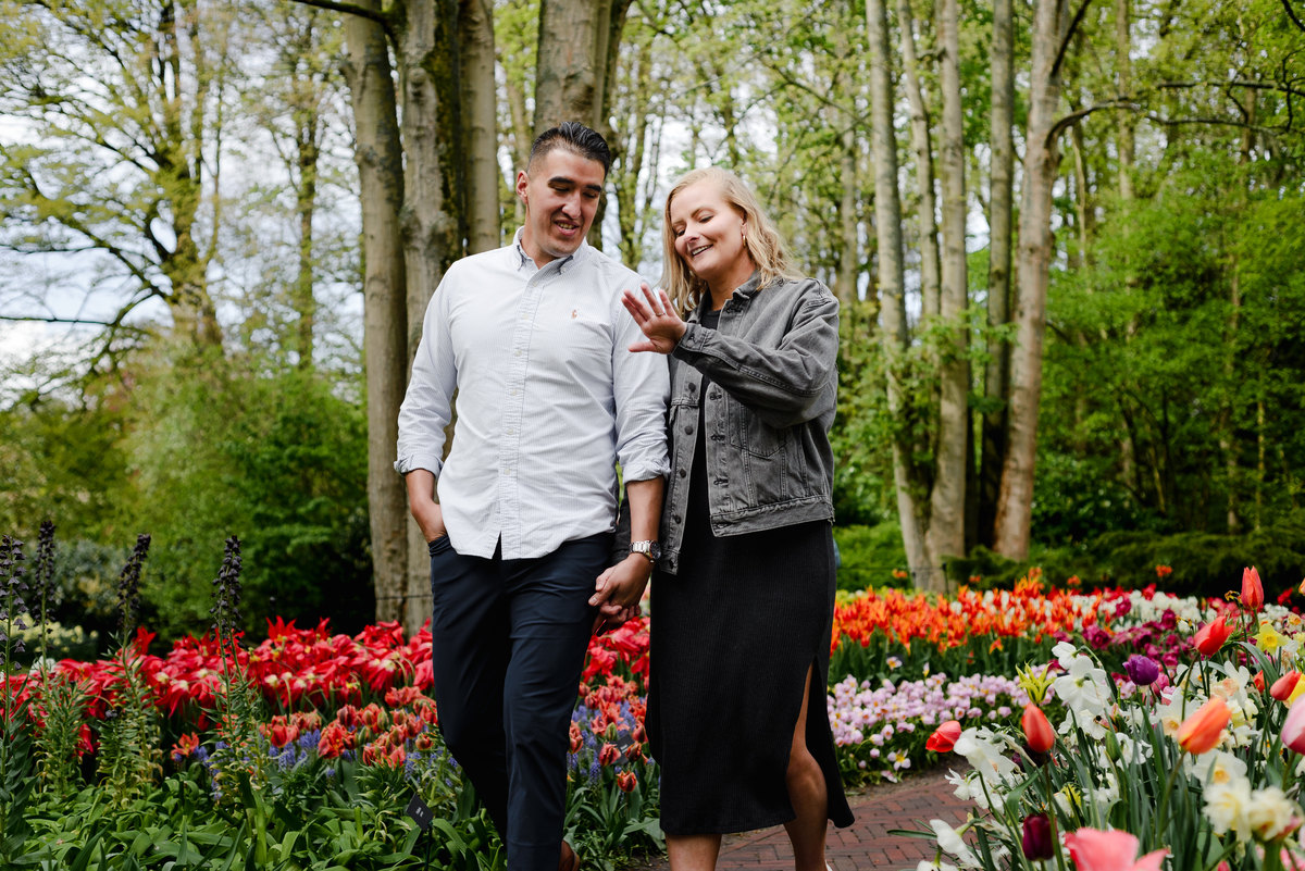 Bride showing engagement ring while walking with partner through Keukenhof tulip gardens, spring flowers and trees behind them.