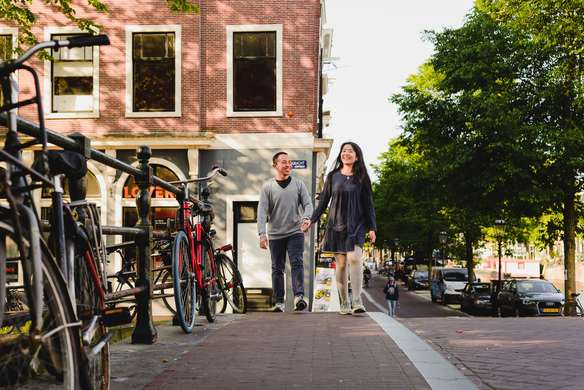 Couple standing close together on an Amsterdam canal bridge with historic buildings and warm evening light.
