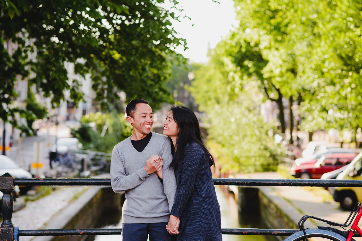 Couple holding hands and smiling on a canal bridge in Amsterdam, surrounded by green trees and water.