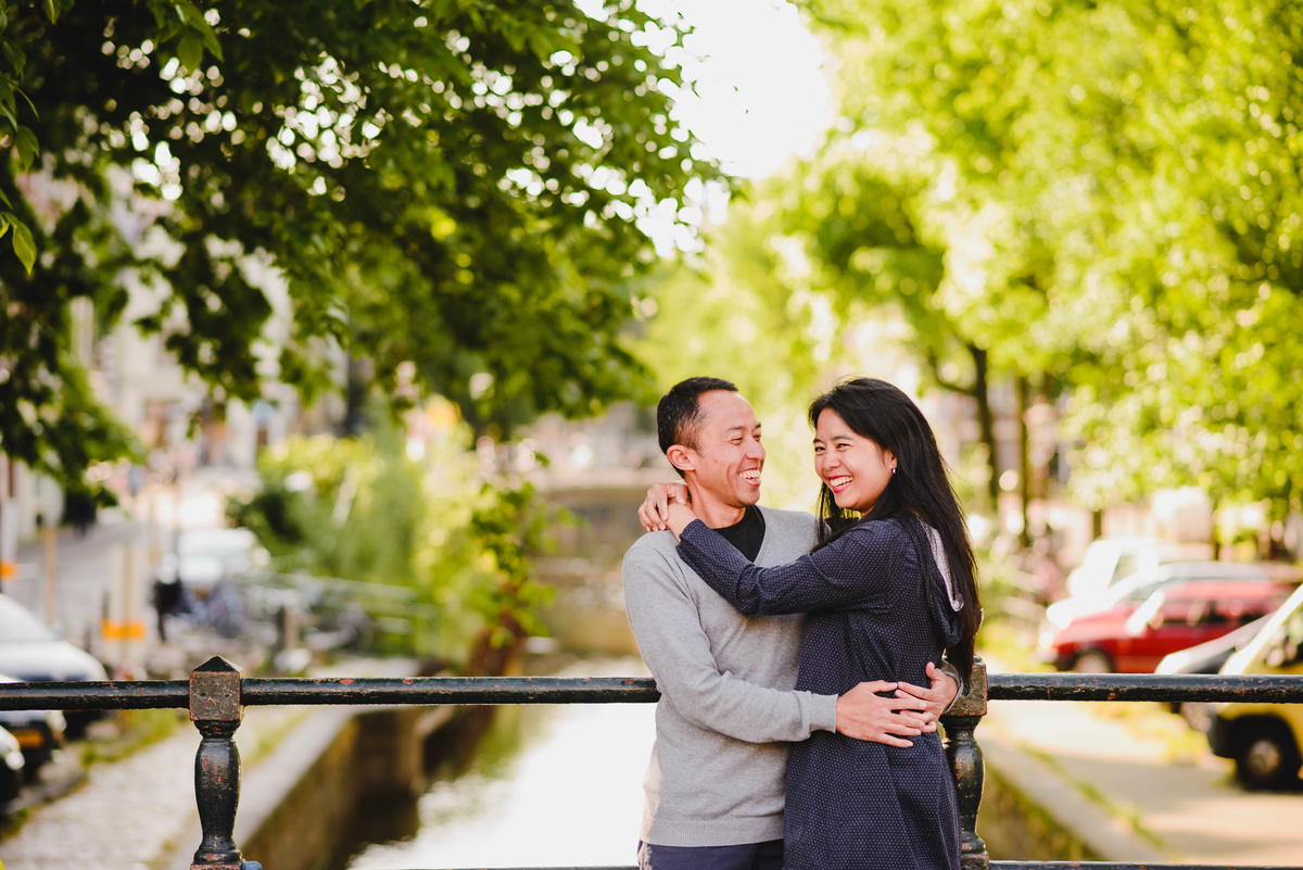Happy couple walking along an Amsterdam canal with historic buildings and trees in the background.