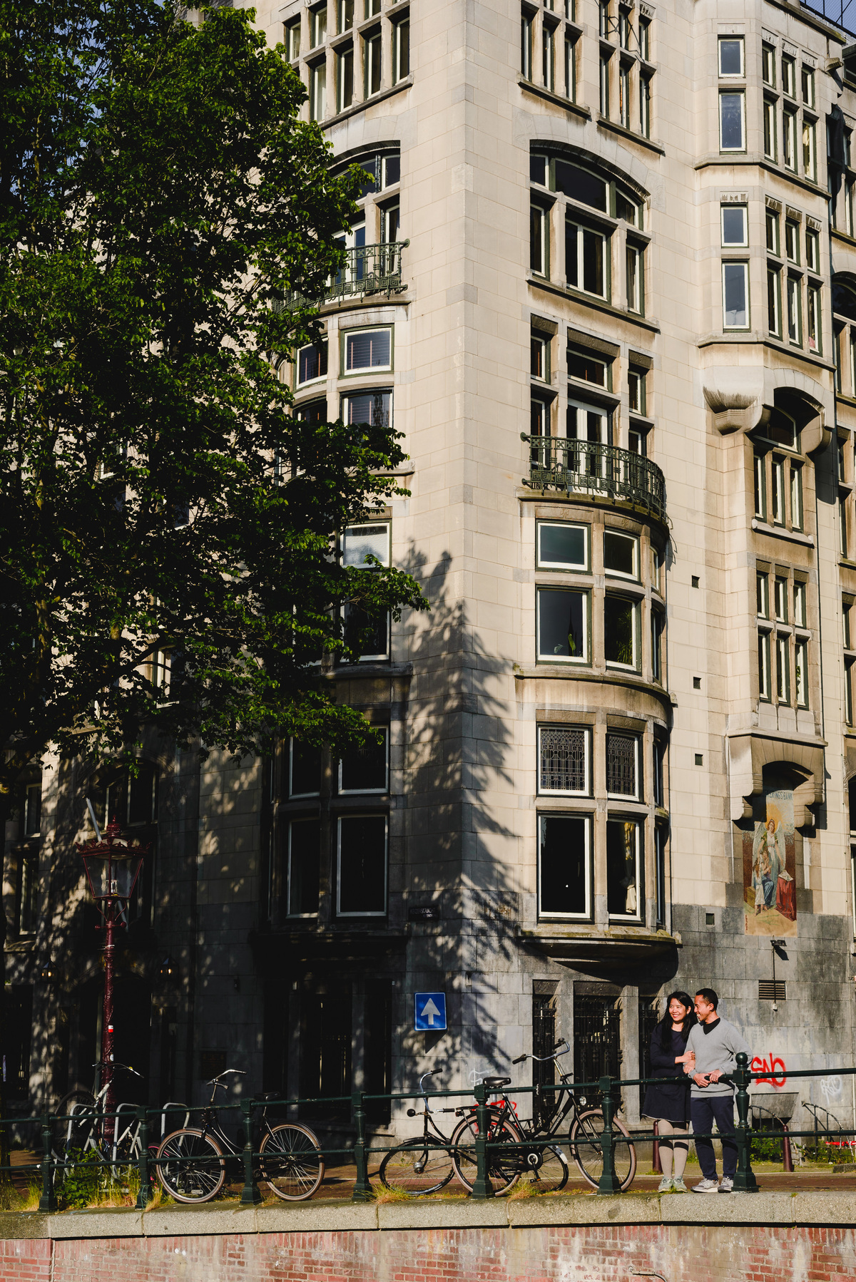 Couple walking together near a canal bridge in Amsterdam, with bicycles and traditional houses around.