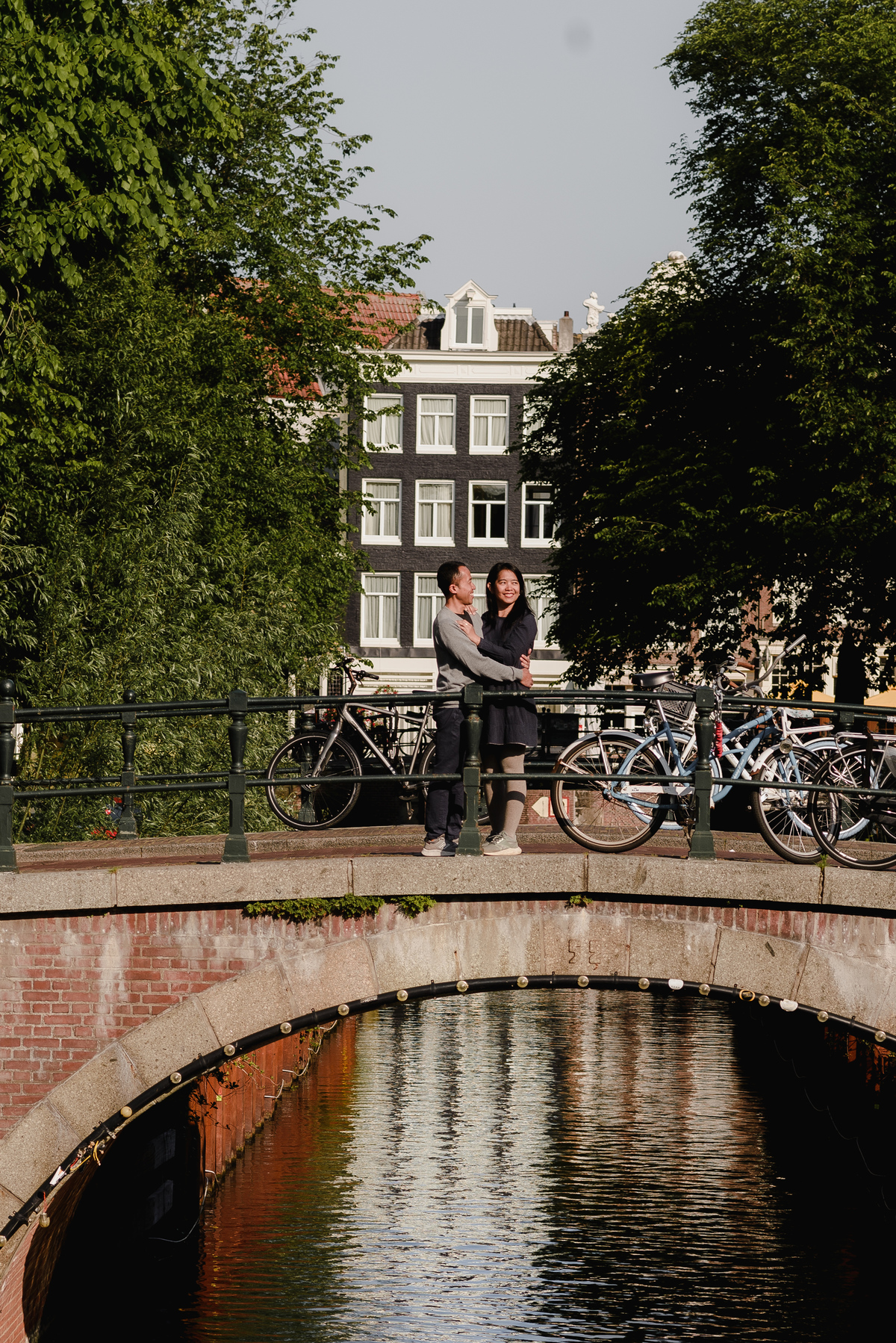 Couple standing on a small canal bridge in Amsterdam, holding each other with bicycles and historic houses in the background