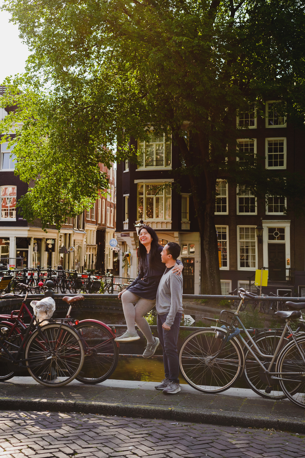 Couple posing together by an Amsterdam canal with bicycles, trees, and warm sunlight during a relaxed photo session.