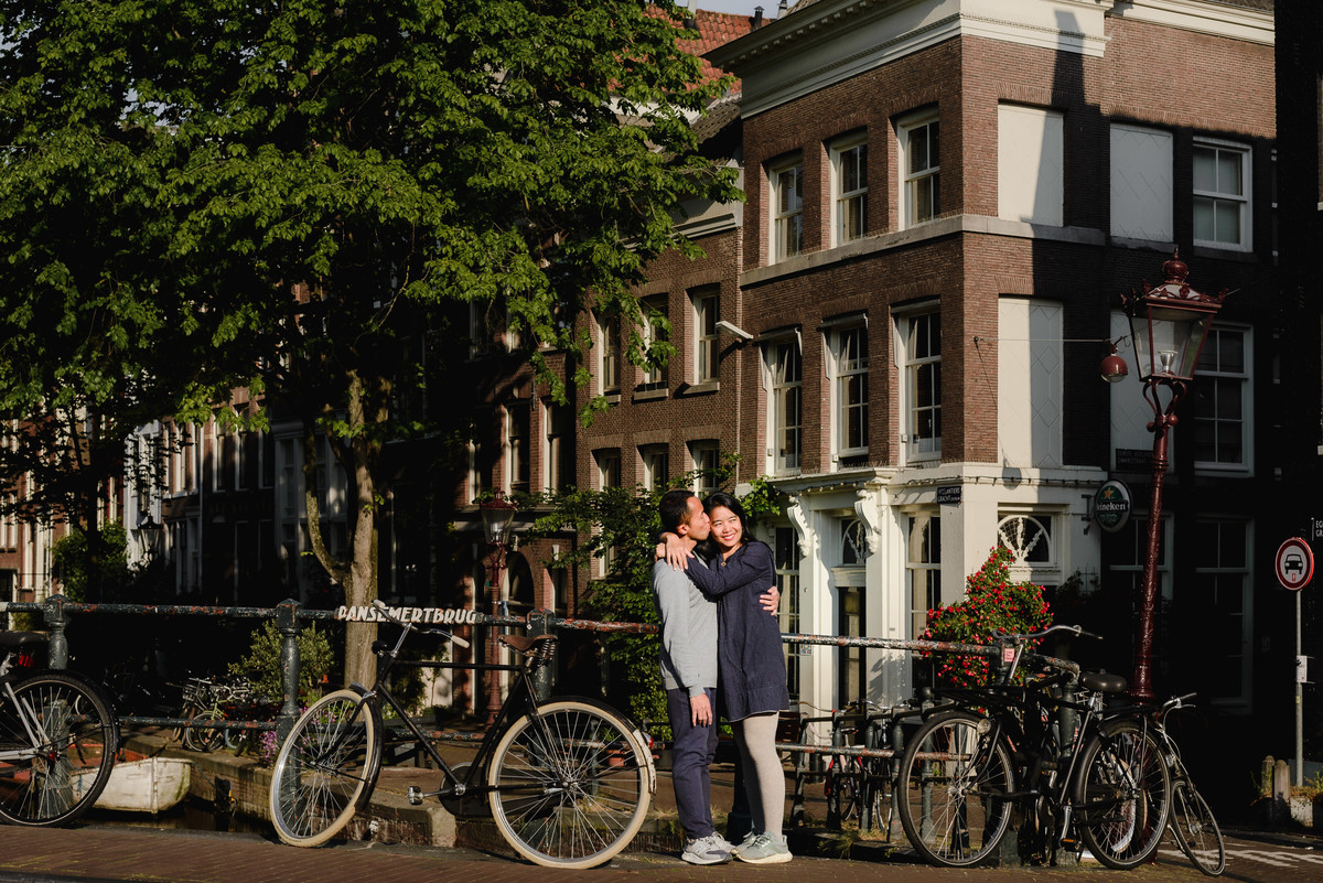 Couple holding hands and walking past a classic Amsterdam corner building on a quiet canal street.