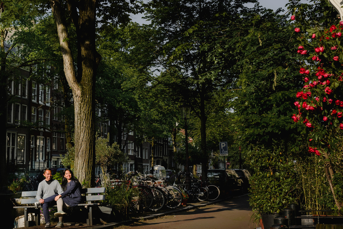 Couple crossing a small Amsterdam street near a canal, framed by historic buildings and soft afternoon light.