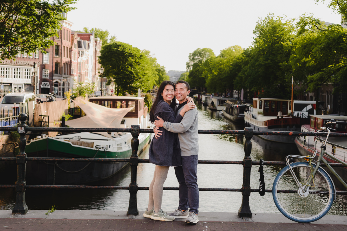Couple embracing on a bridge over an Amsterdam canal with houseboats and trees in the background.