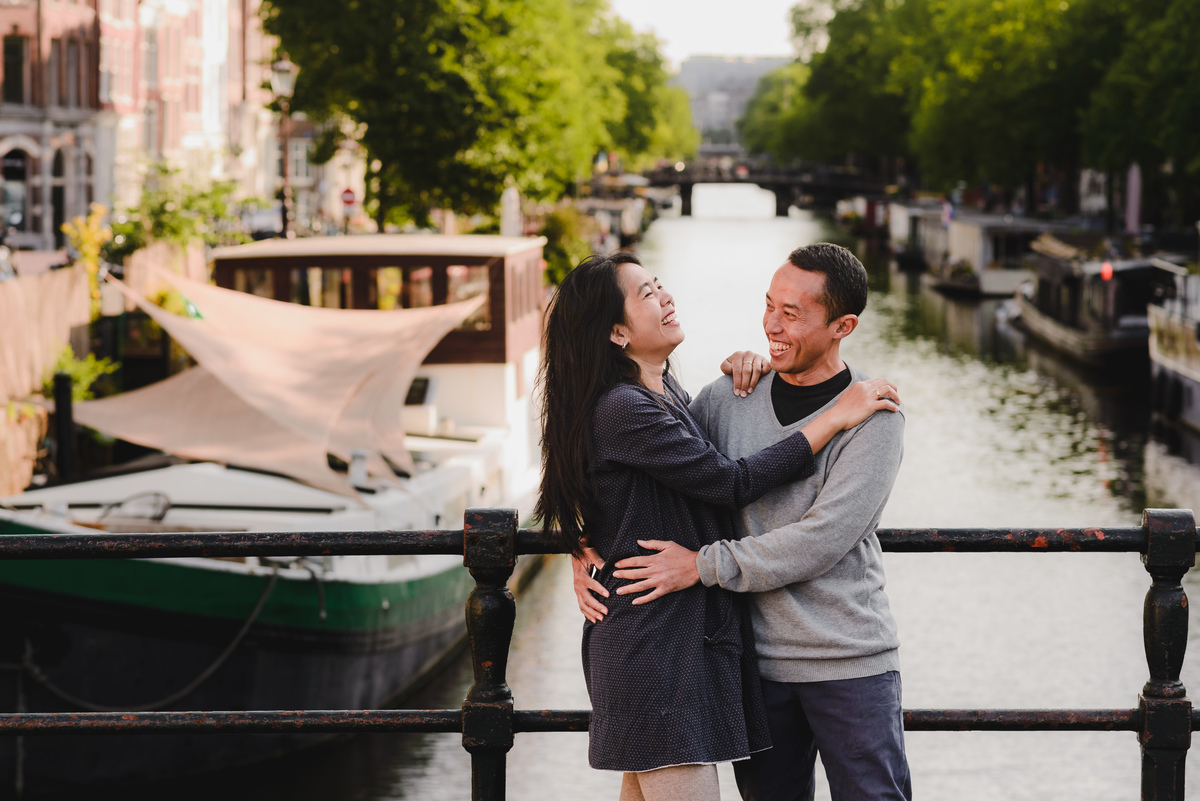 Smiling couple hugging on an Amsterdam canal bridge with boats and green trees reflecting on the water.