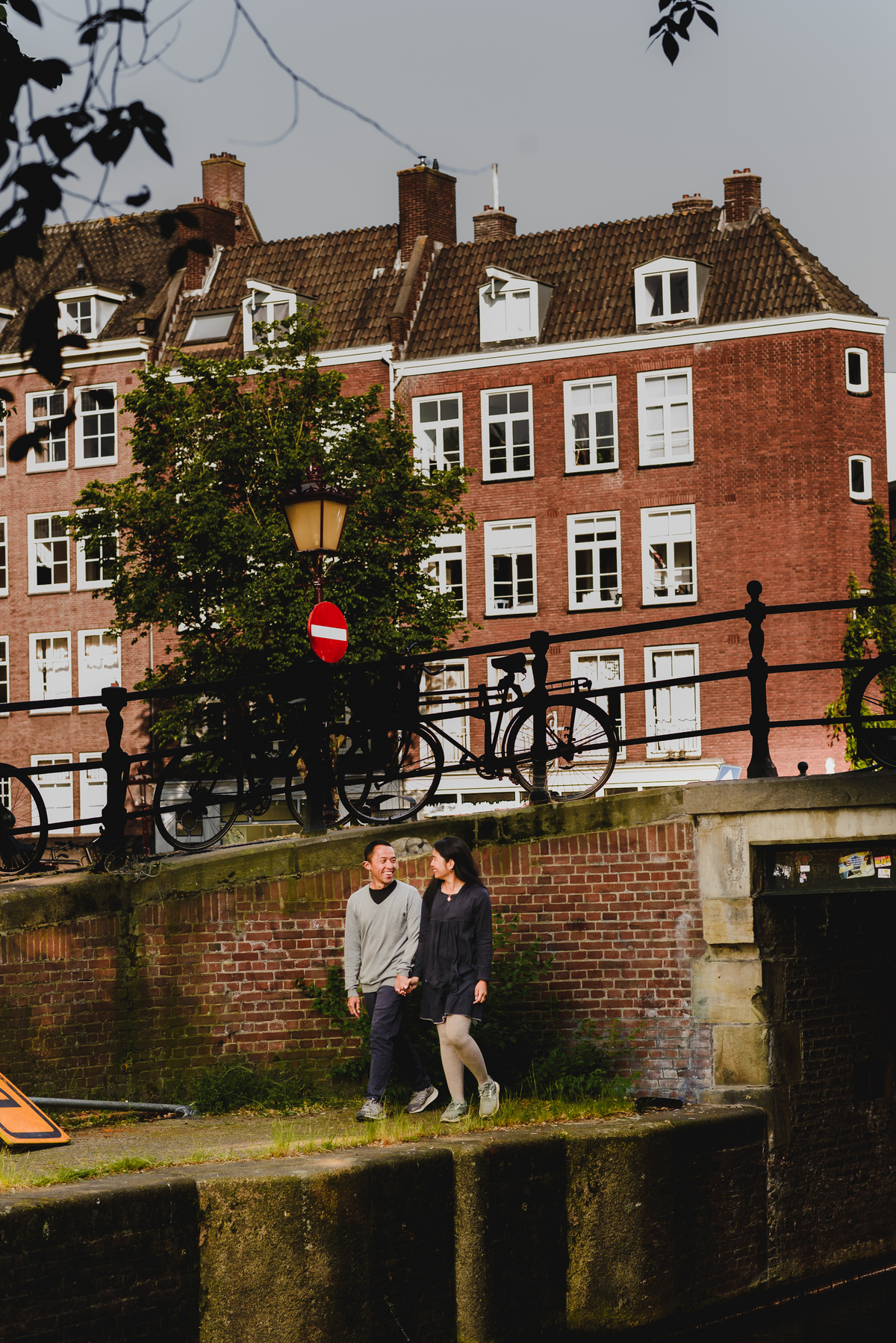Couple walking hand in hand past a traditional Amsterdam café and historic canal houses.