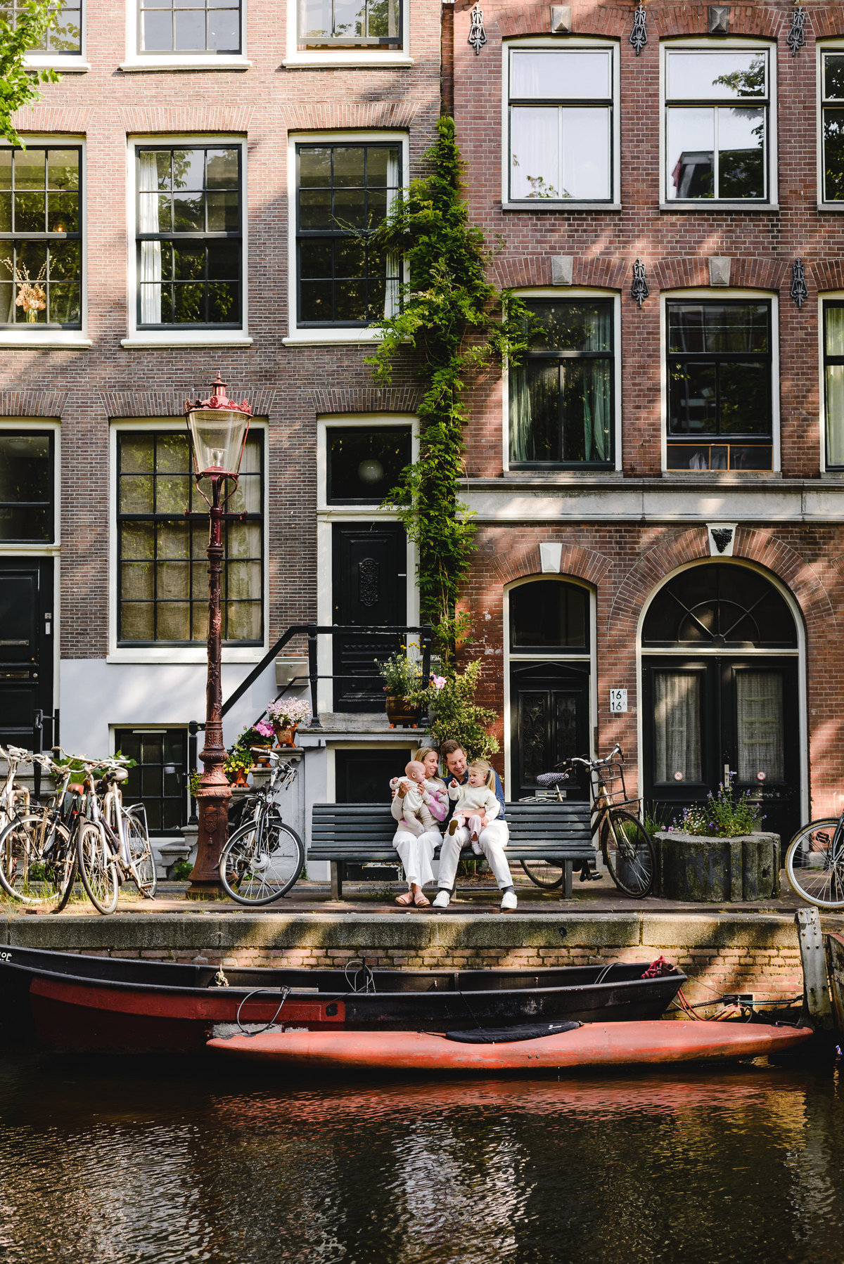 Family laughing together on a canal bench in Amsterdam, with bicycles and brick canal houses behind them.