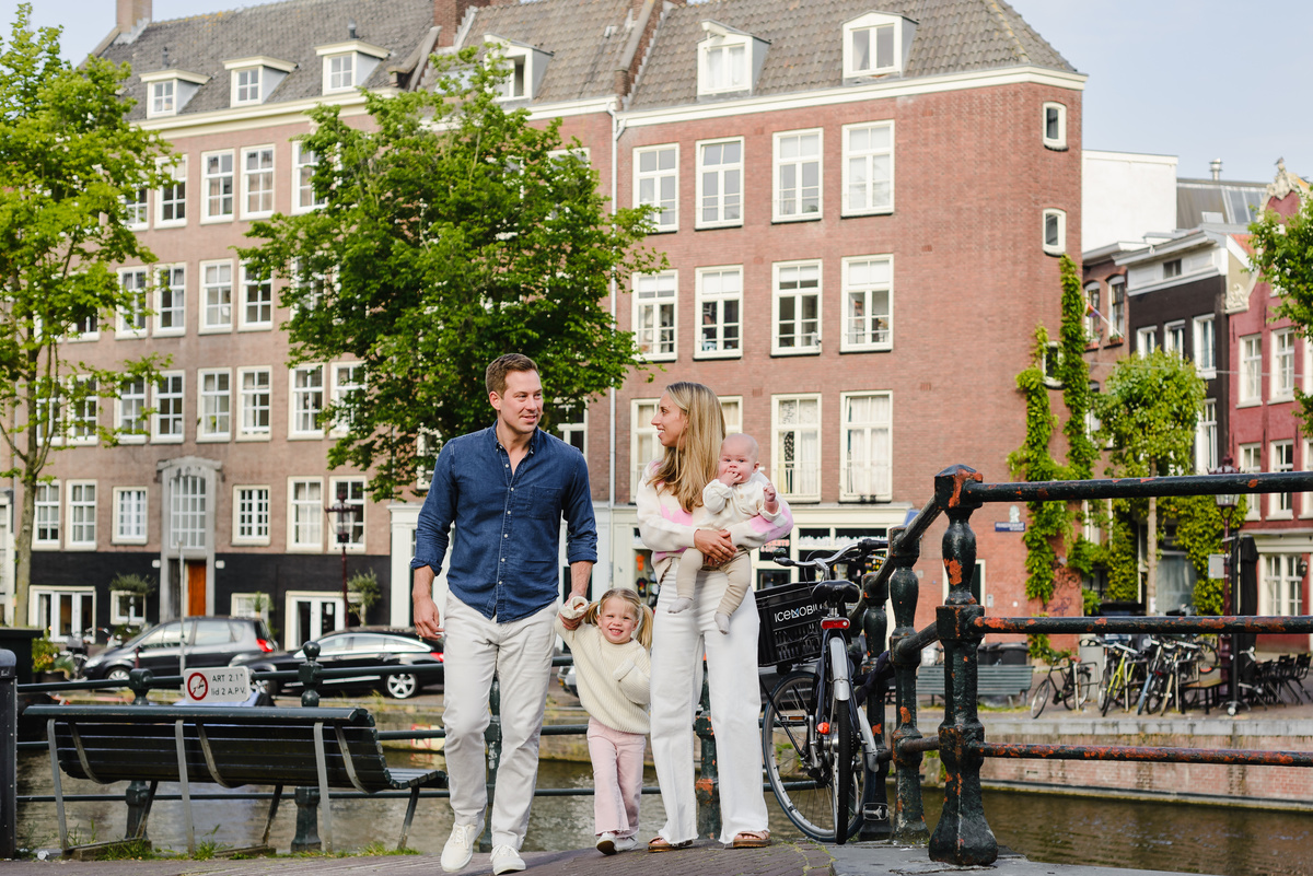 Family smiling together on a canal bridge in Amsterdam, parents holding their children with boats and greenery in the background.