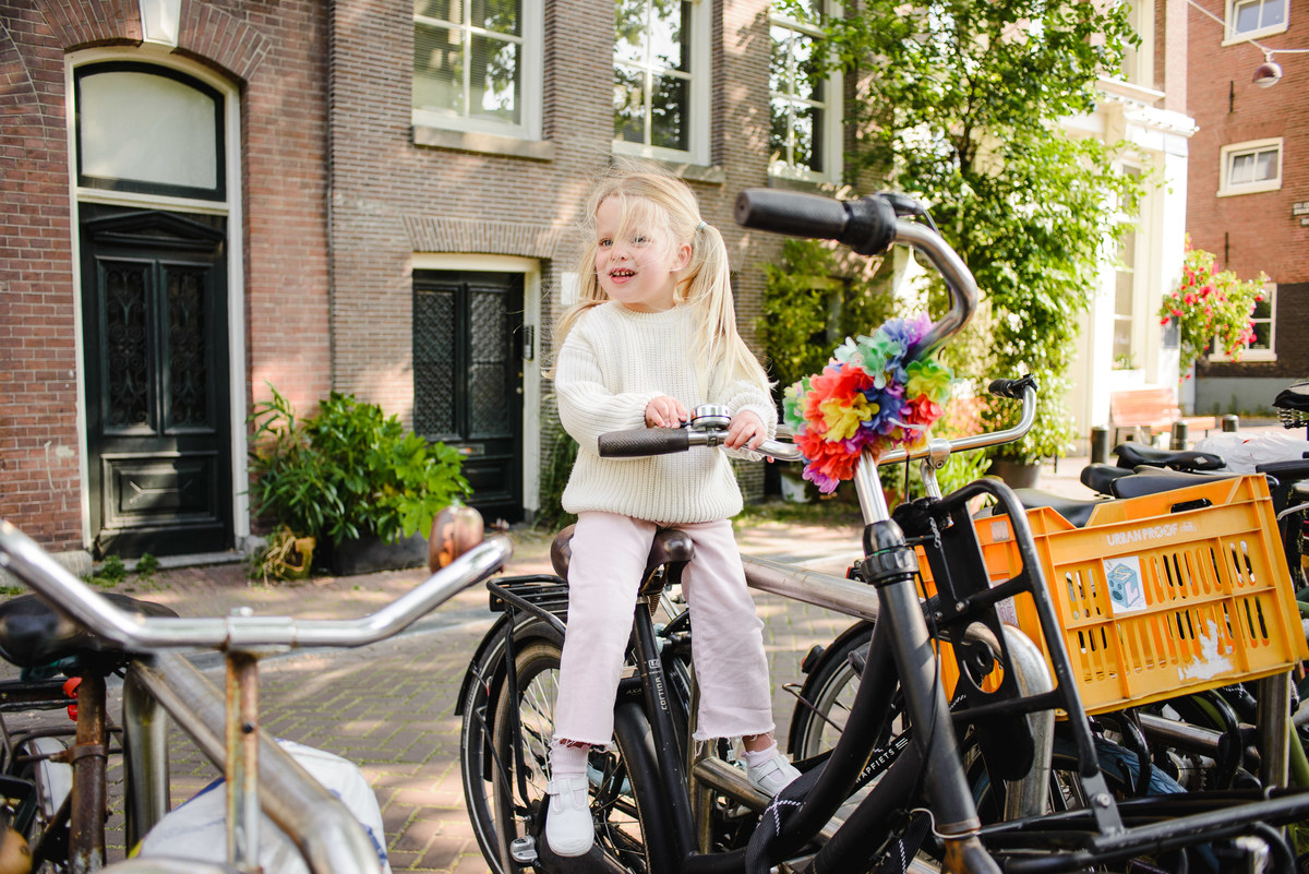 Toddler sitting on a bicycle seat, looking at the camera, with Amsterdam street, bikes, and canal houses in the background.